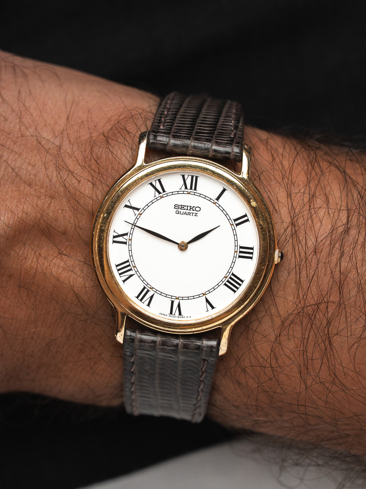 Montre Seiko Quartz vintage au boîtier doré, cadran blanc et bracelet en cuir gris.