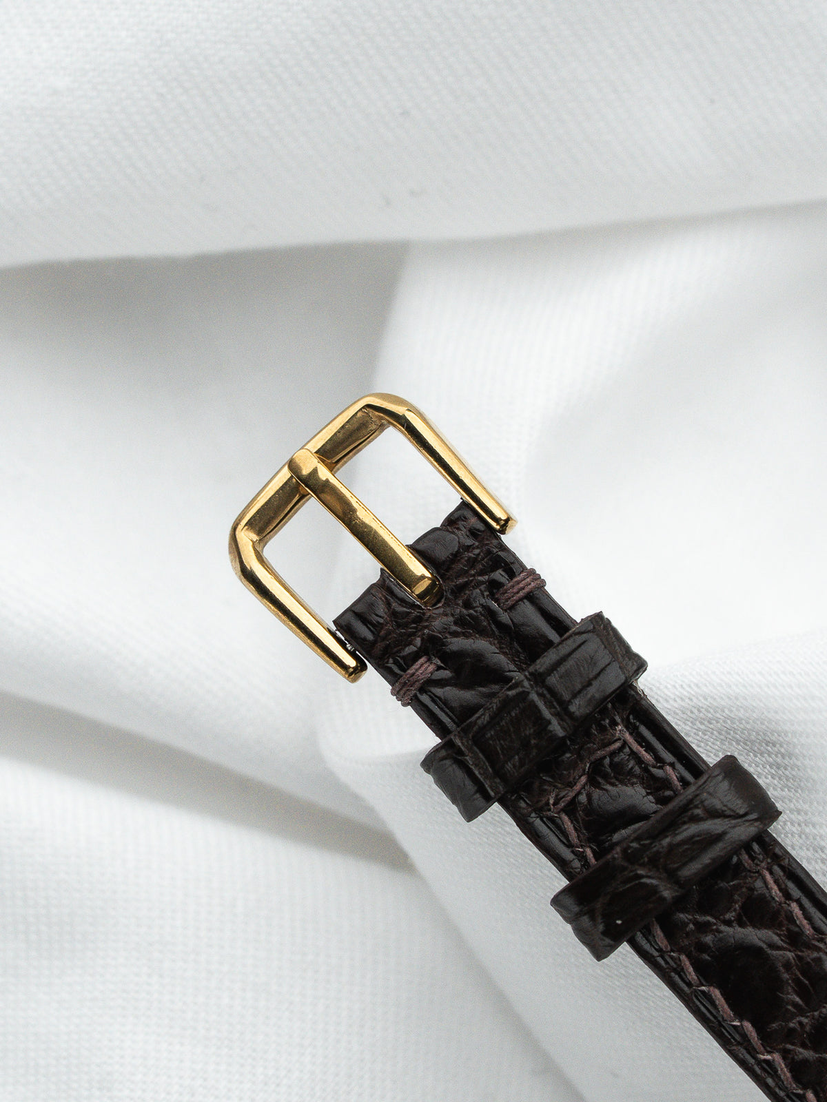 Boucle dorée et bracelet en cuir marron façon crocodile sur fond de tissu blanc.