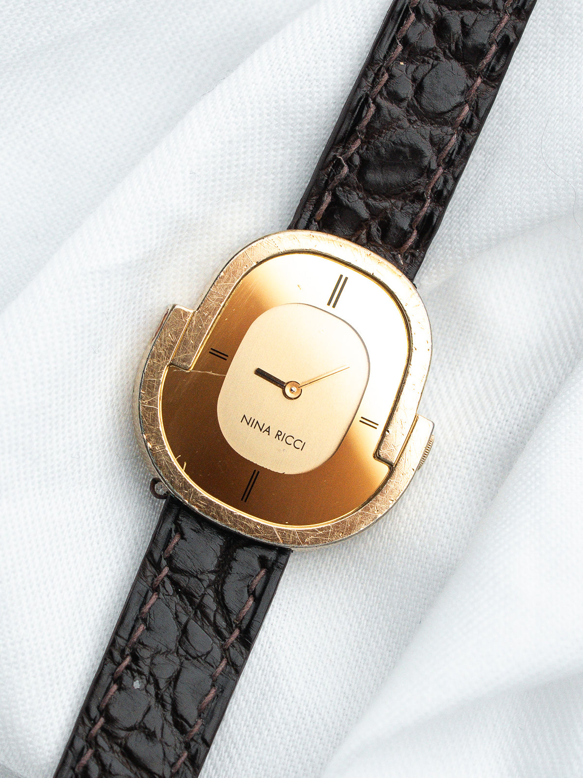 Montre Nina Ricci vintage dorée avec cadran ovale et bracelet en cuir marron texturé.