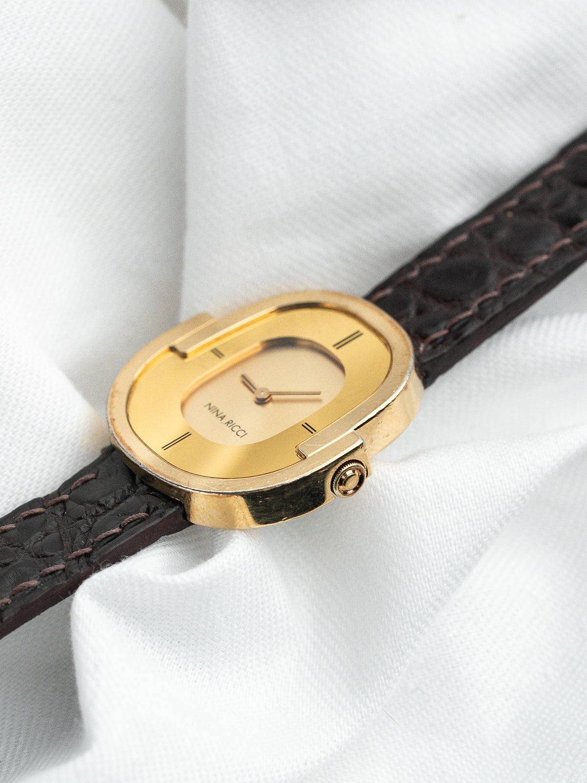 Montre Nina Ricci vintage en or avec bracelet en cuir marron sur tissu blanc.