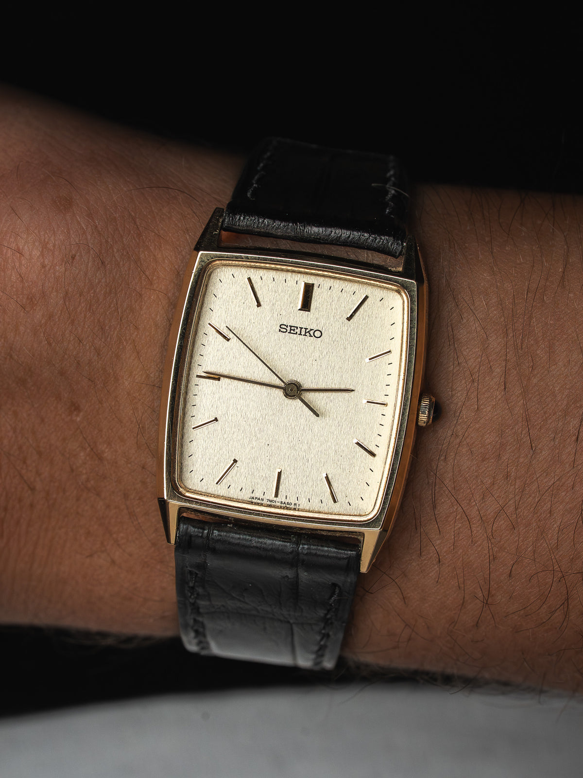 Montre Seiko vintage au boîtier rectangulaire doré avec cadran clair et bracelet en cuir noir.