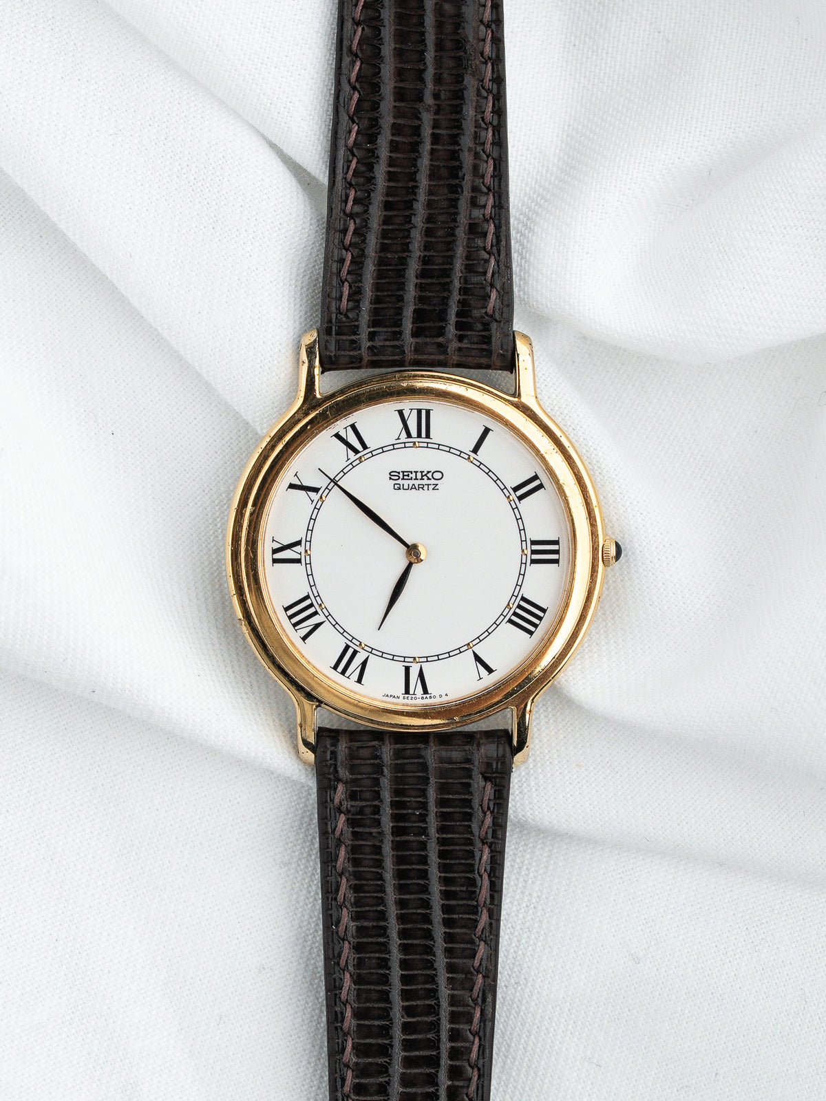 Montre Seiko Quartz vintage au boîtier doré, cadran blanc et bracelet en cuir brun.