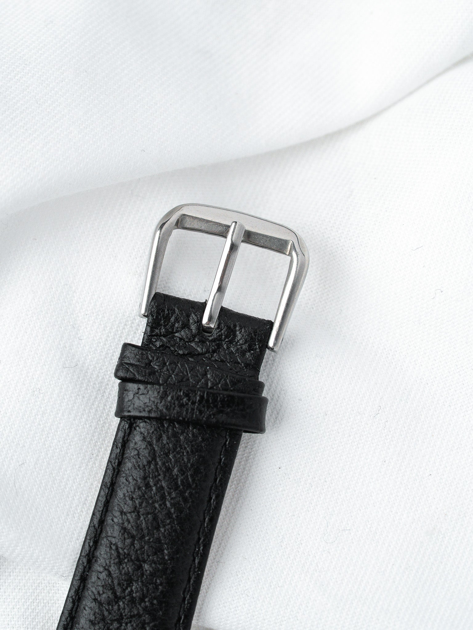 Black textured leather watch strap with silver buckle on white fabric