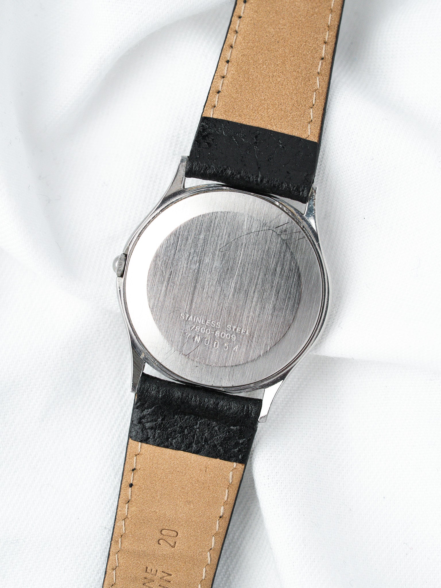 Vintage stainless steel watch back with engraved serial number, black leather strap on white fabric