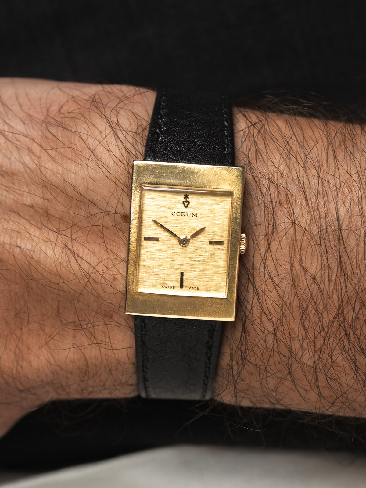 Montre Corum vintage au boîtier carré doré et bracelet en cuir noir, portée au poignet