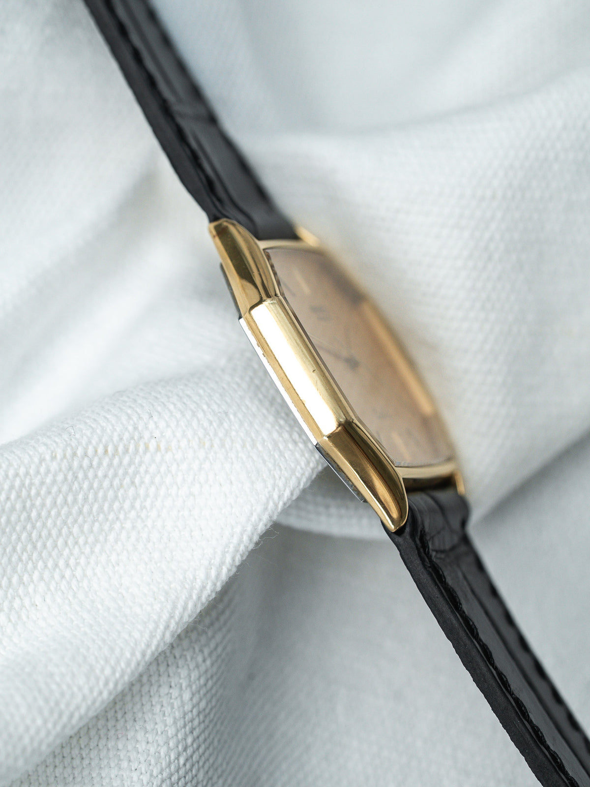 Montre vintage au boîtier doré rectangulaire avec bracelet en cuir noir sur tissu blanc.
