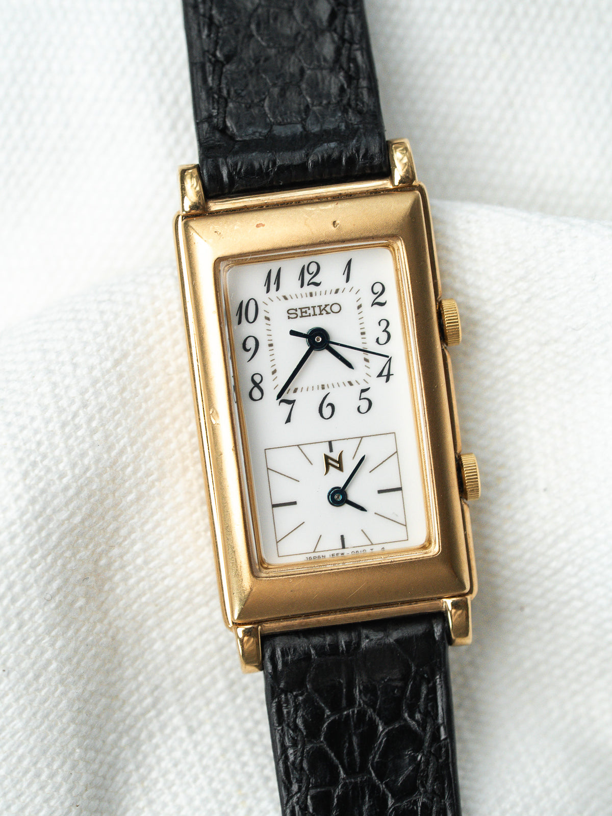 Montre Seiko vintage rectangulaire en or avec cadran blanc et bracelet cuir noir.