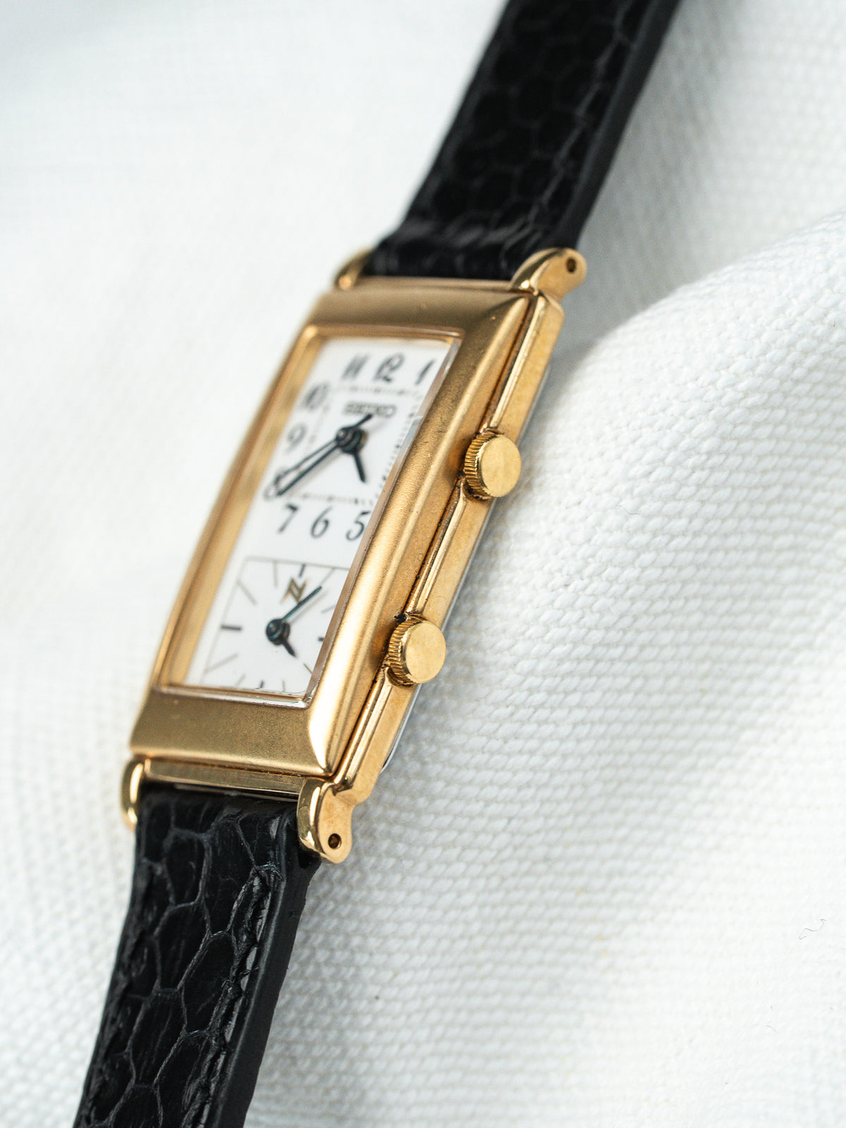 Montre rectangulaire dorée avec cadran blanc et bracelet en cuir noir texturé.