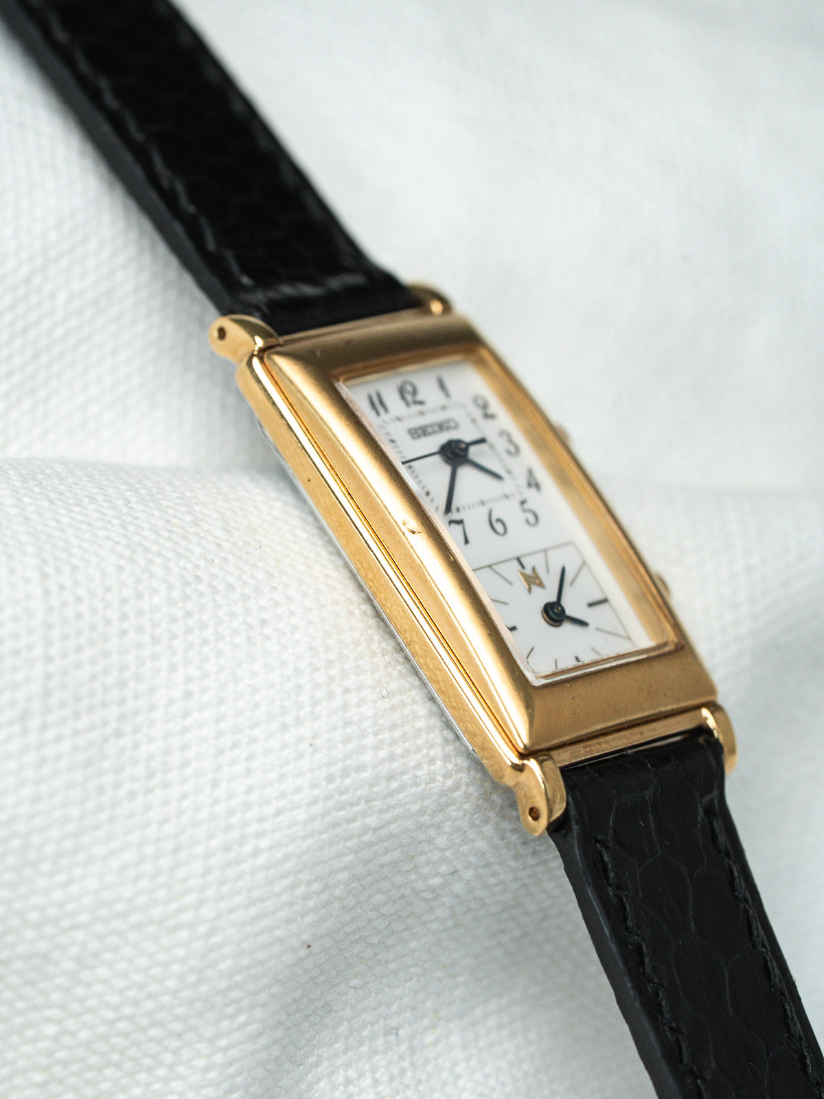 Montre vintage Seiko à boîtier rectangulaire doré, cadran blanc et bracelet en cuir noir.