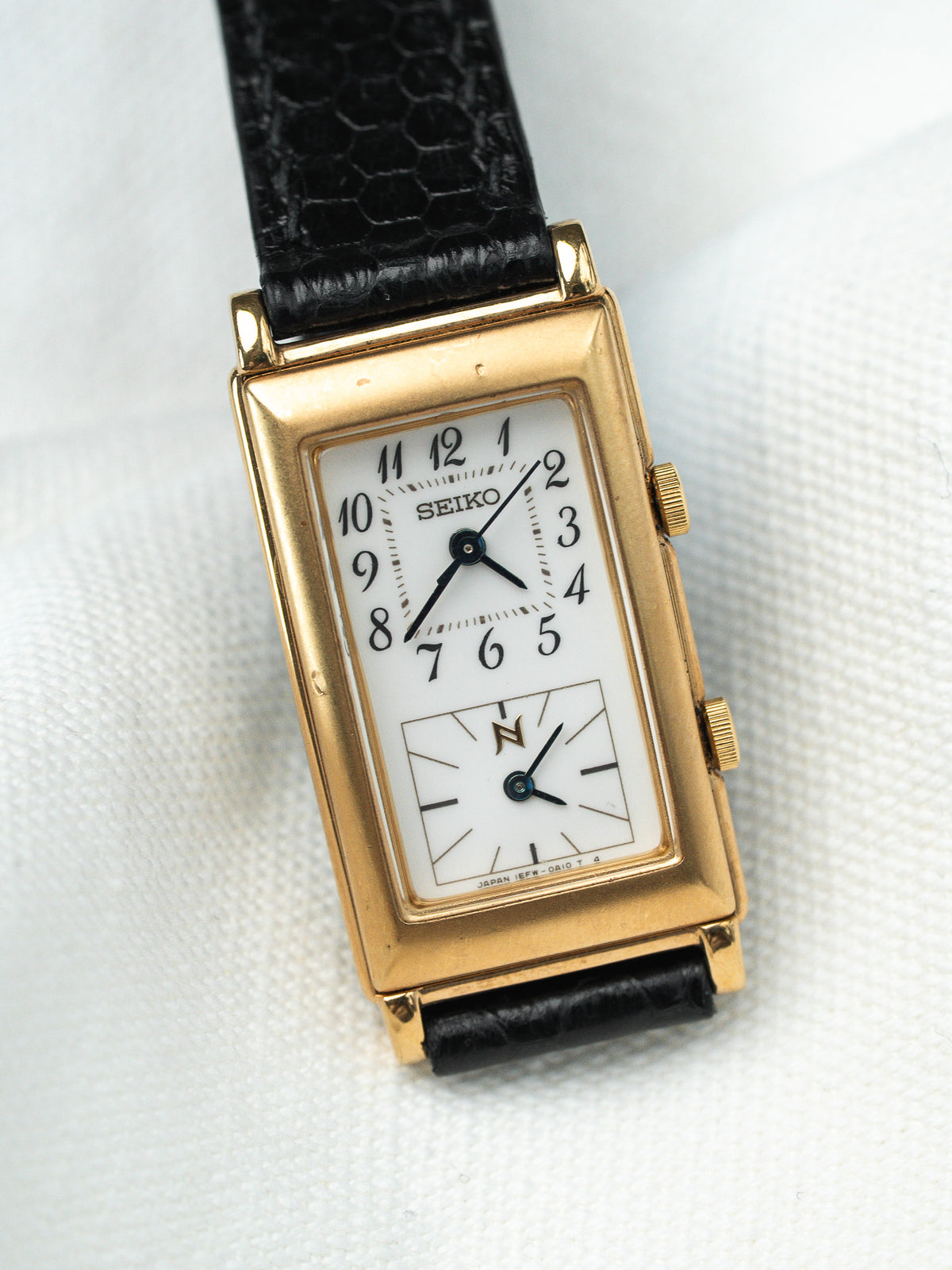 Montre Seiko vintage à boîtier rectangulaire doré, cadran blanc et bracelet en cuir noir.