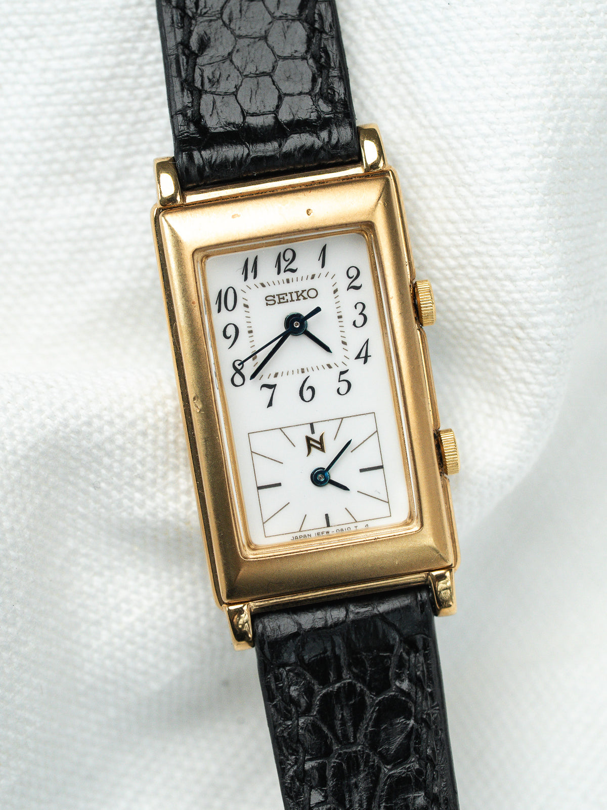 Montre Seiko vintage rectangulaire dorée avec cadran blanc et bracelet en cuir noir.