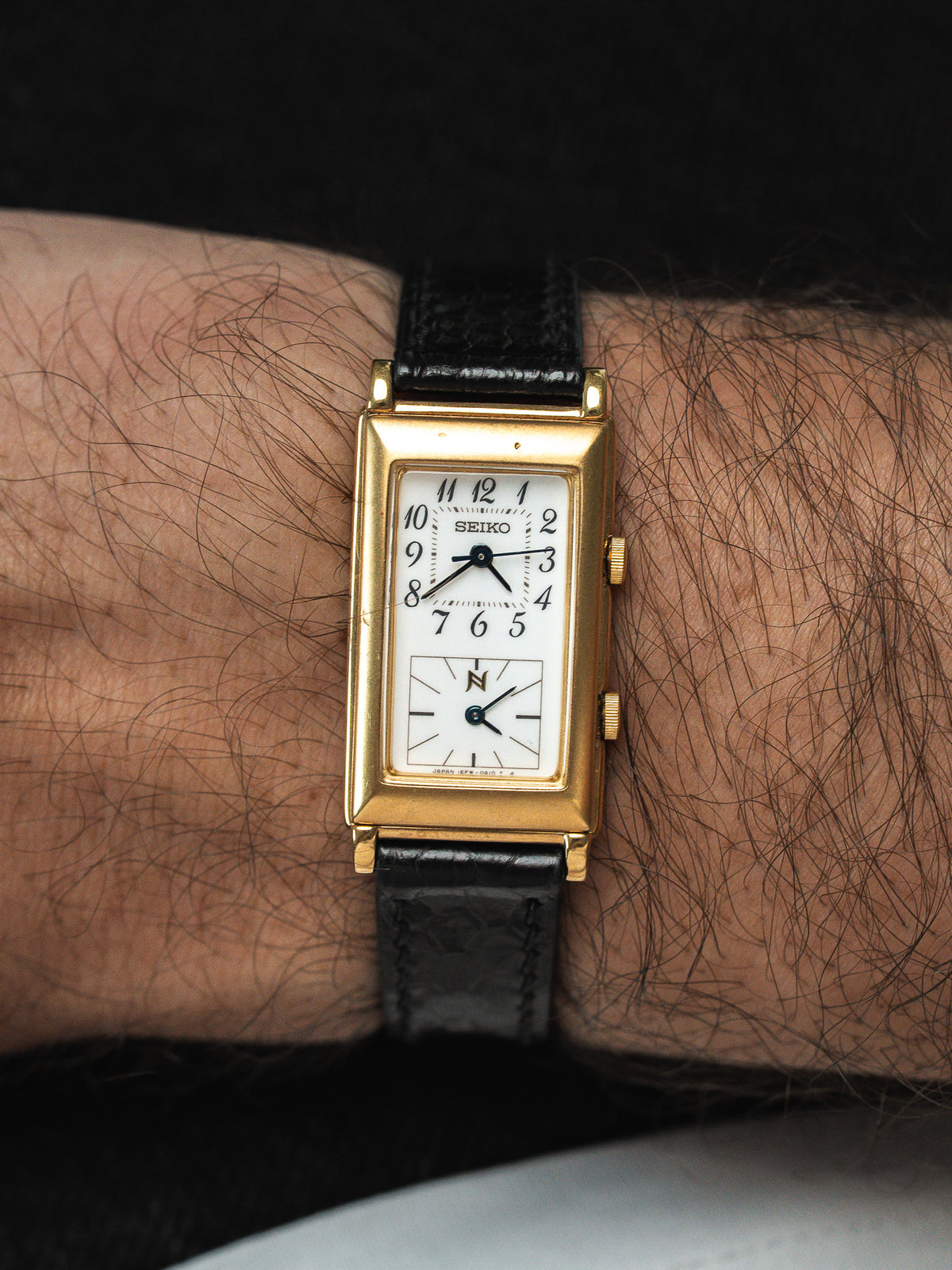 Montre Seiko vintage rectangulaire en or avec cadran blanc et bracelet cuir noir.