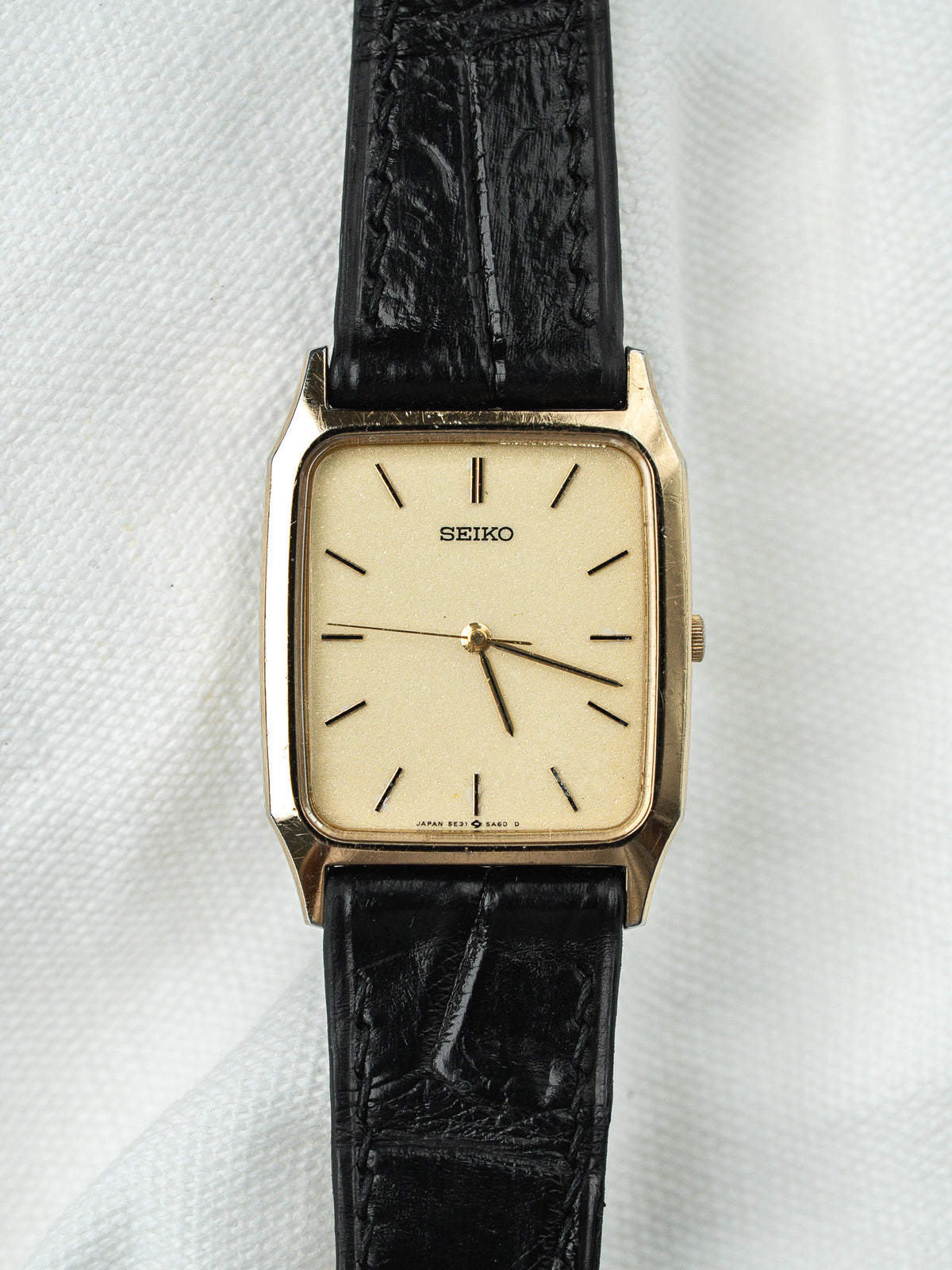 Montre Seiko vintage rectangulaire dorée avec cadran champagne et bracelet en cuir noir.