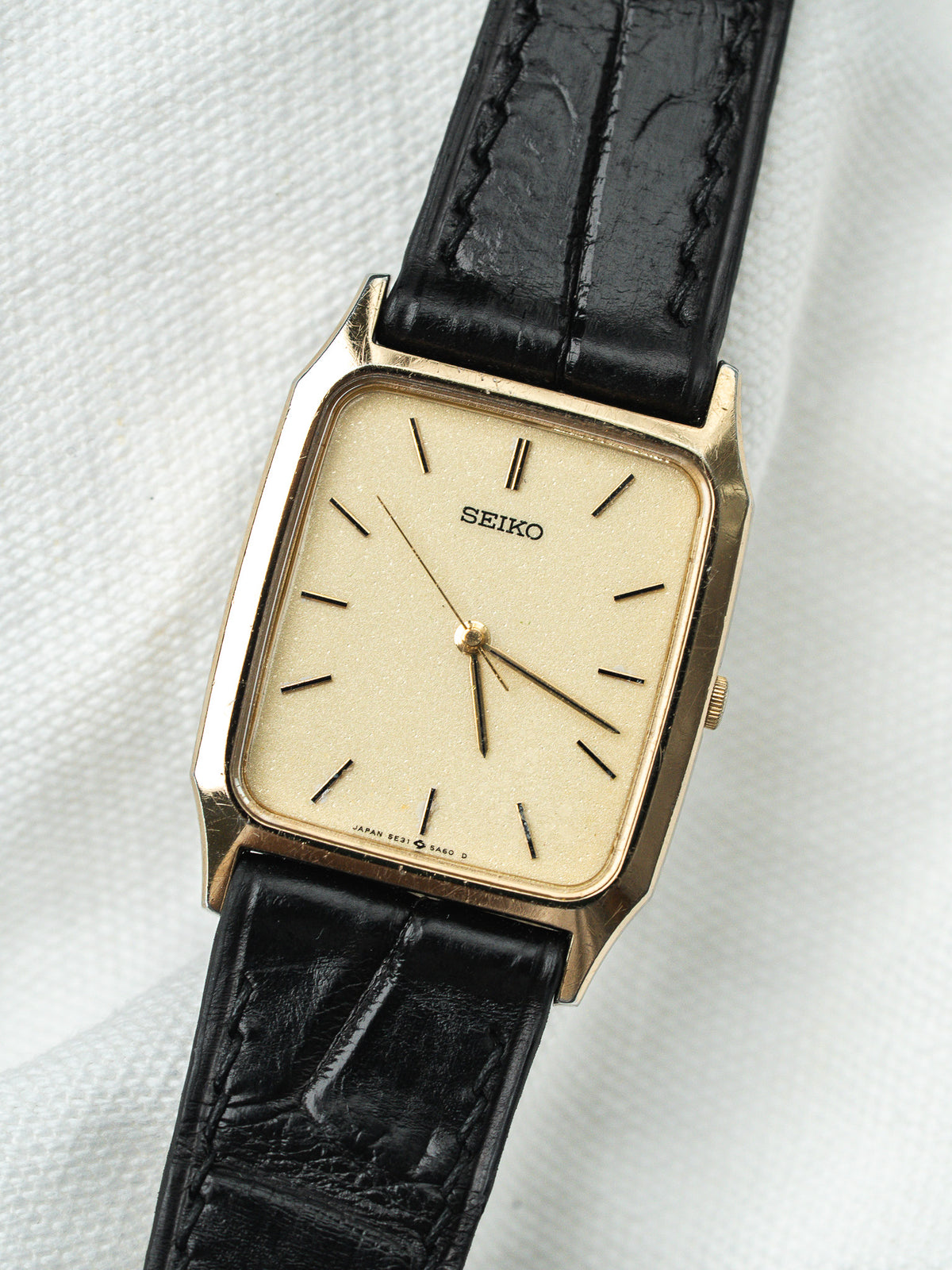 Montre Seiko vintage avec boîtier rectangulaire doré, cadran beige et bracelet en cuir noir.
