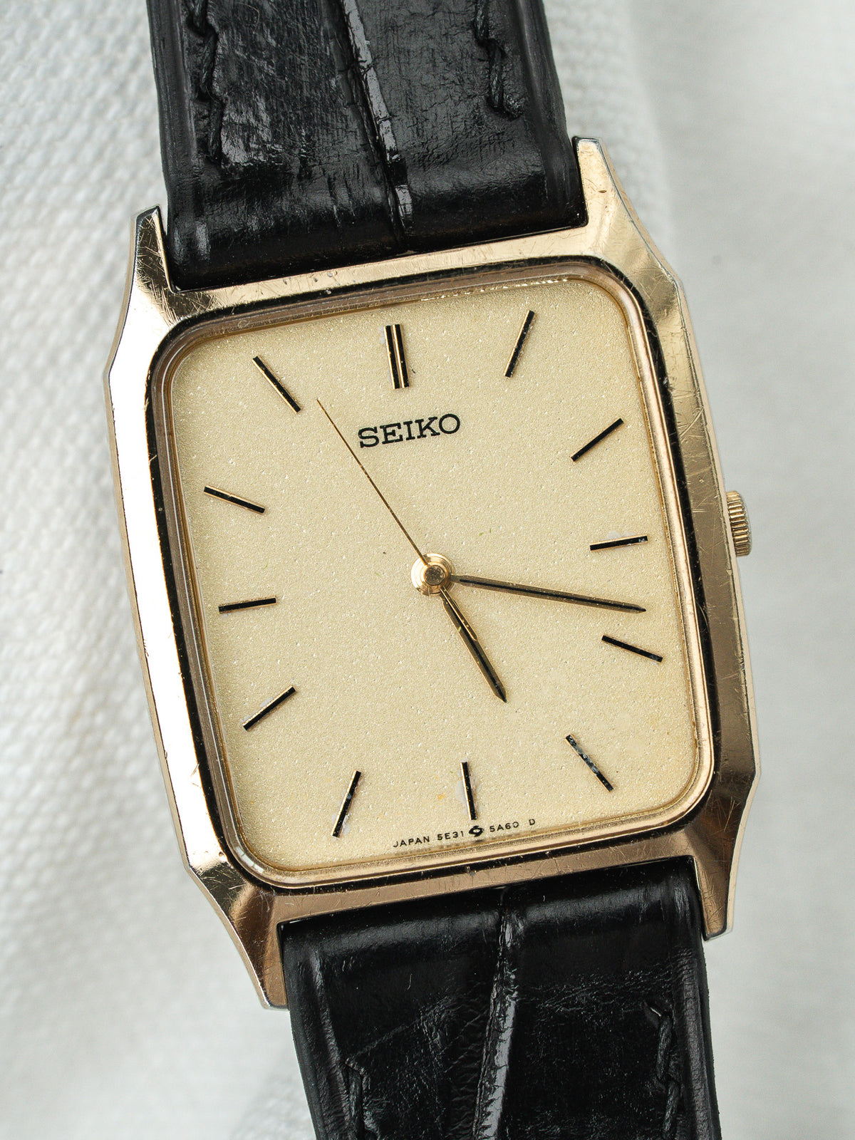 Montre Seiko vintage à boîtier rectangulaire doré, cadran crème et bracelet en cuir noir.