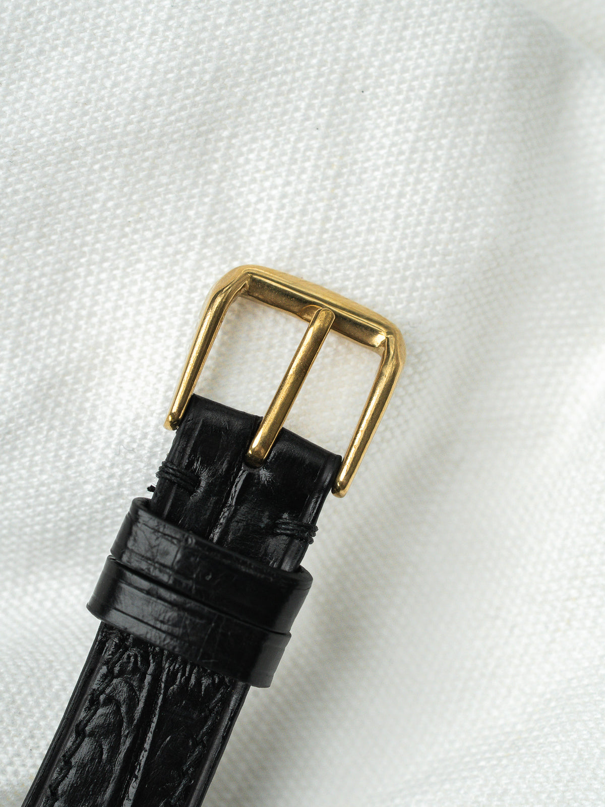 Boucle de montre dorée sur un bracelet en cuir noir grainé sur fond blanc.