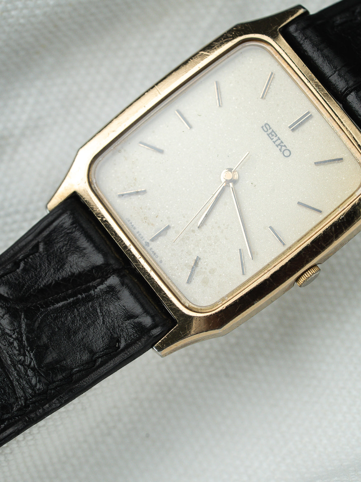 Montre Seiko vintage avec boîtier doré rectangulaire, cadran blanc et bracelet en cuir noir.