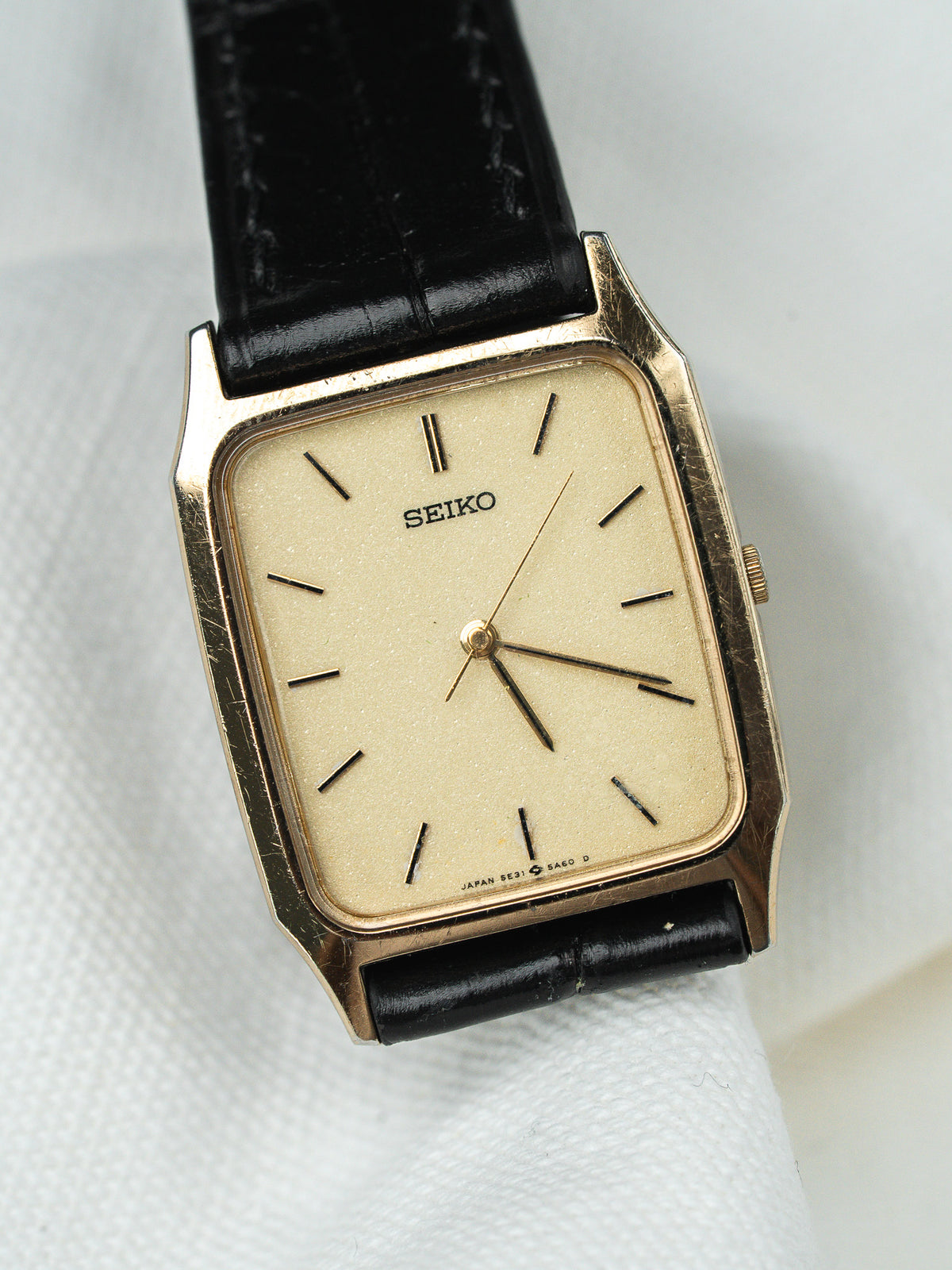Montre vintage Seiko à boîtier rectangulaire doré, cadran champagne et bracelet en cuir noir.