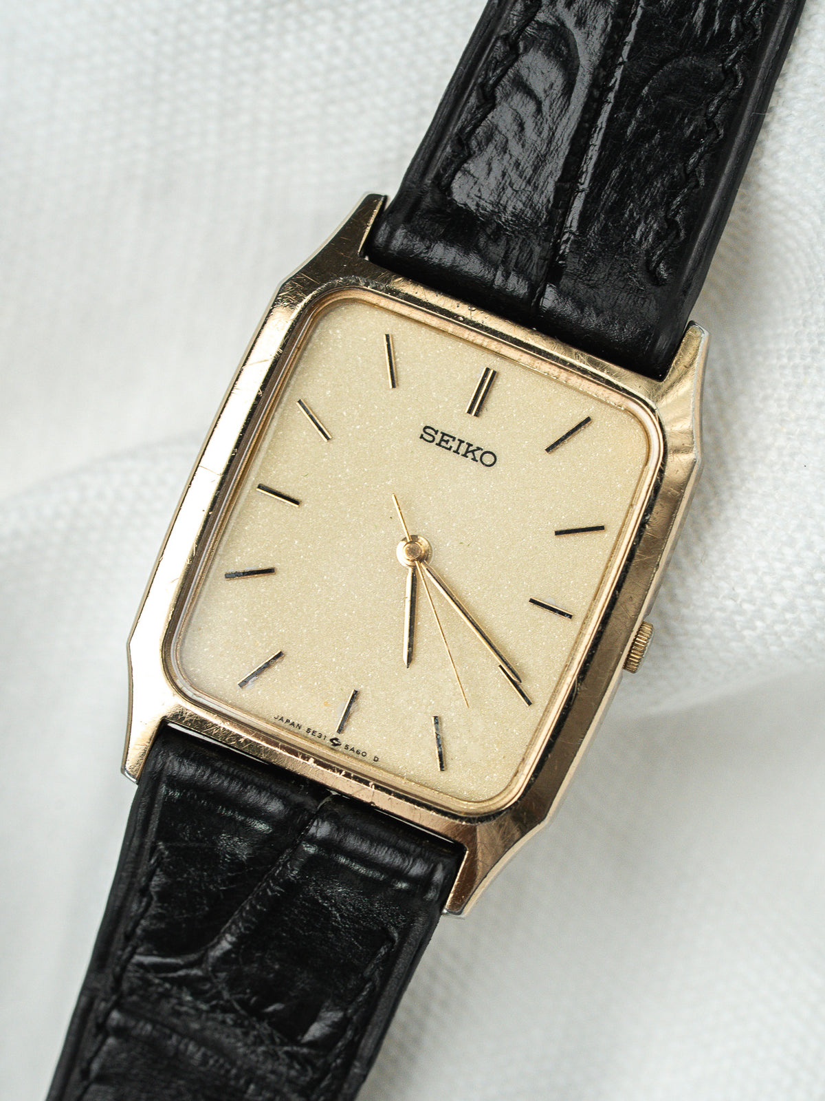 Montre Seiko vintage avec boîtier doré rectangulaire, cadran beige et bracelet en cuir noir.