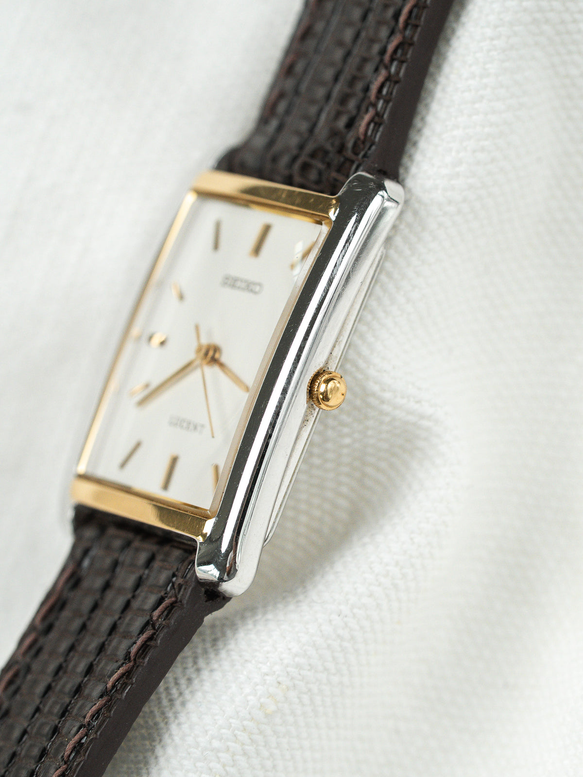 Montre vintage rectangulaire avec cadran blanc, boîtier doré et bracelet en cuir brun.