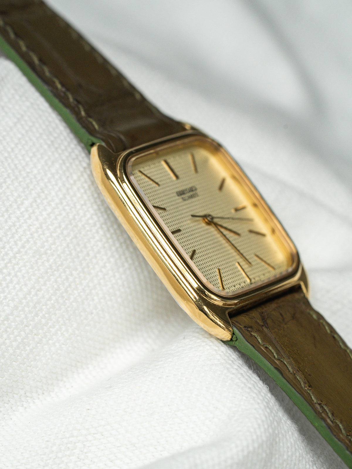 Montre vintage à boîtier rectangulaire doré avec cadran texturé et bracelet en cuir marron.