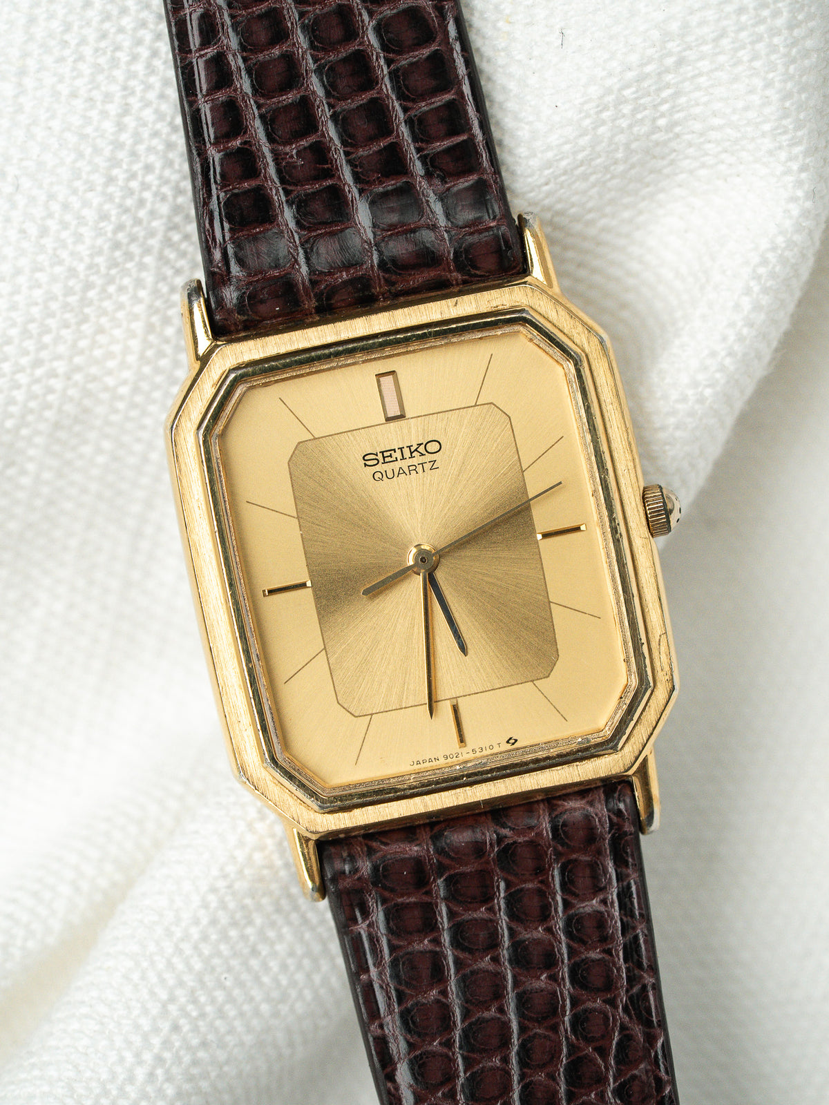 Montre Seiko Quartz vintage au boîtier doré rectangulaire et bracelet en cuir bordeaux.