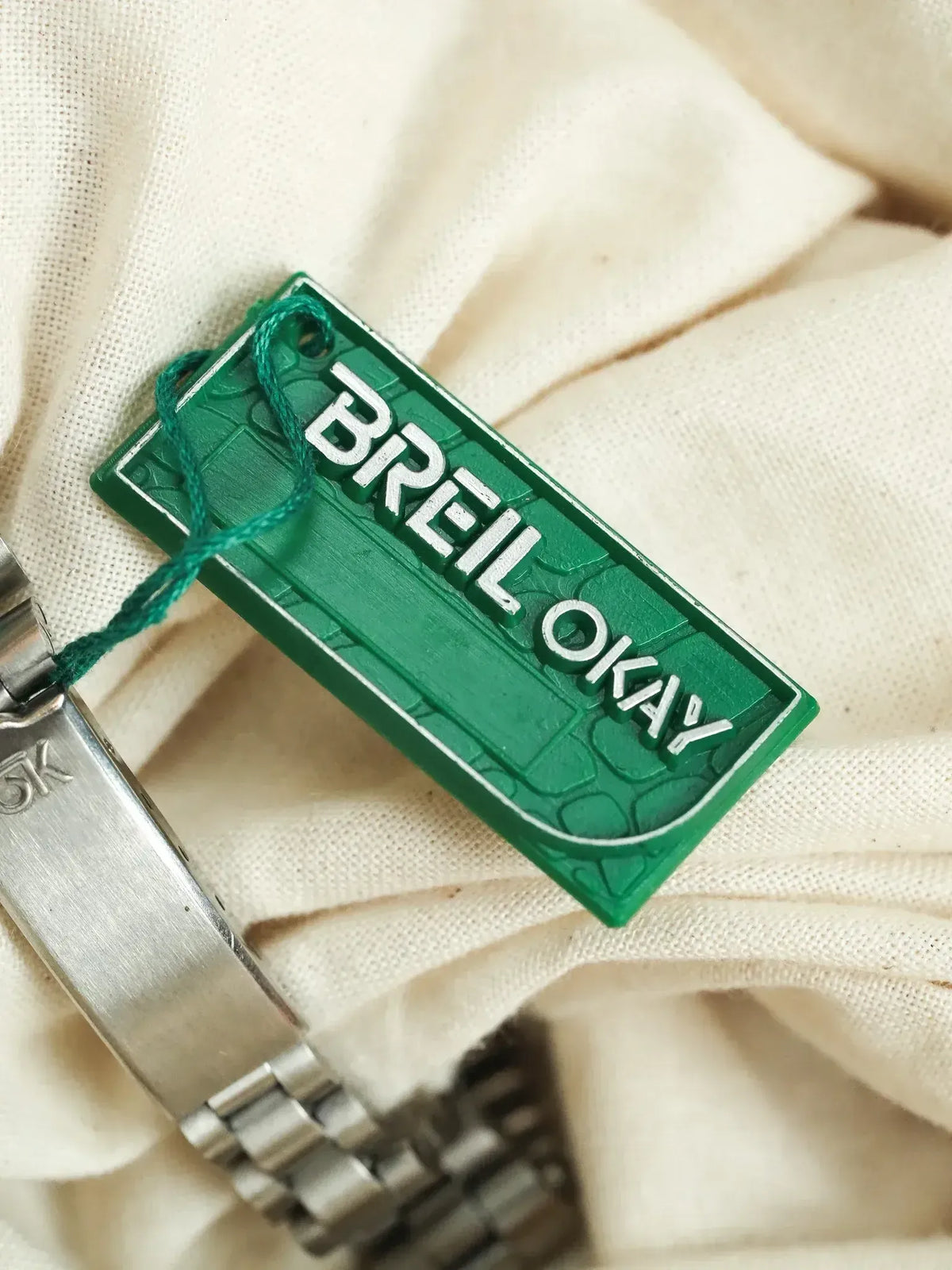 BREL OKAY green tag attached to stainless steel watch bracelet on fabric