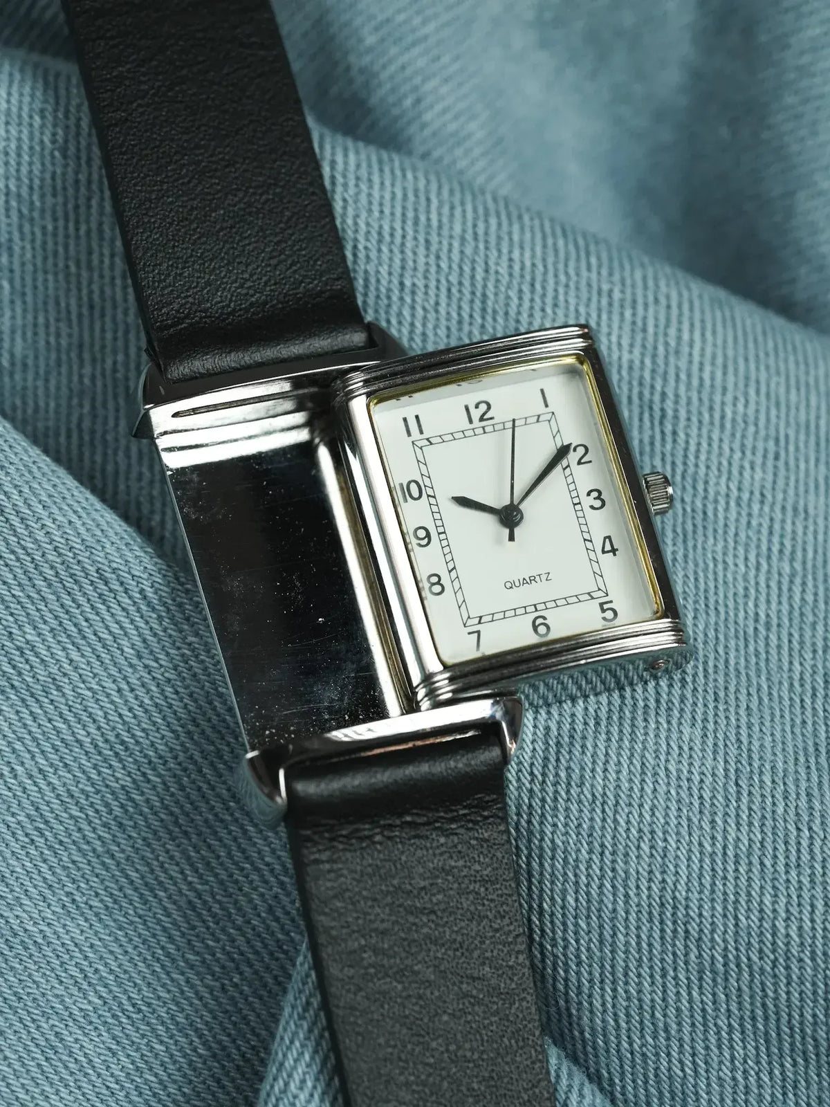 Vintage quartz watch with rectangular silver case, white dial, black leather strap on blue fabric
