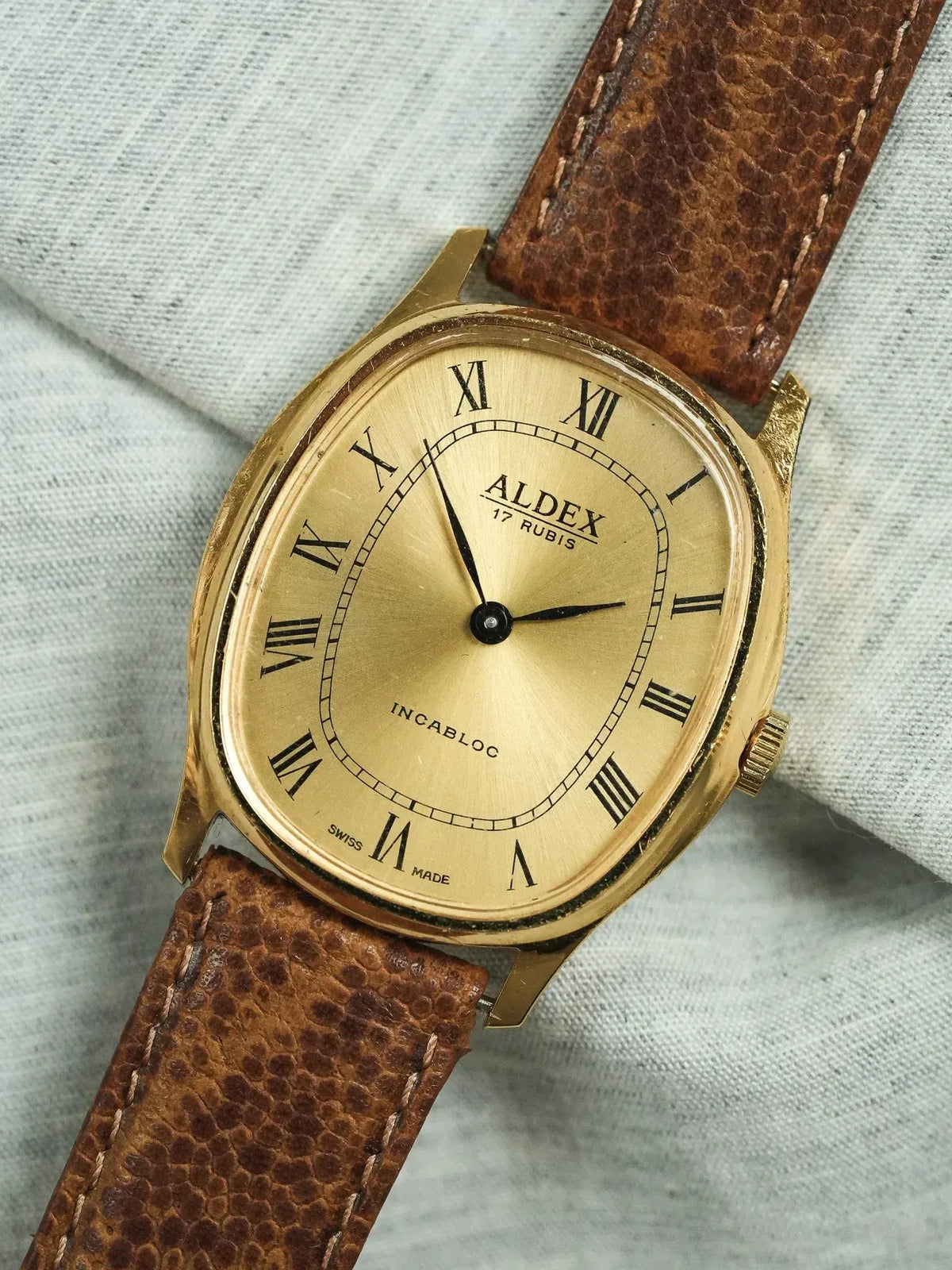 Vintage Aldex watch with gold case, Roman numerals, brown leather strap on fabric