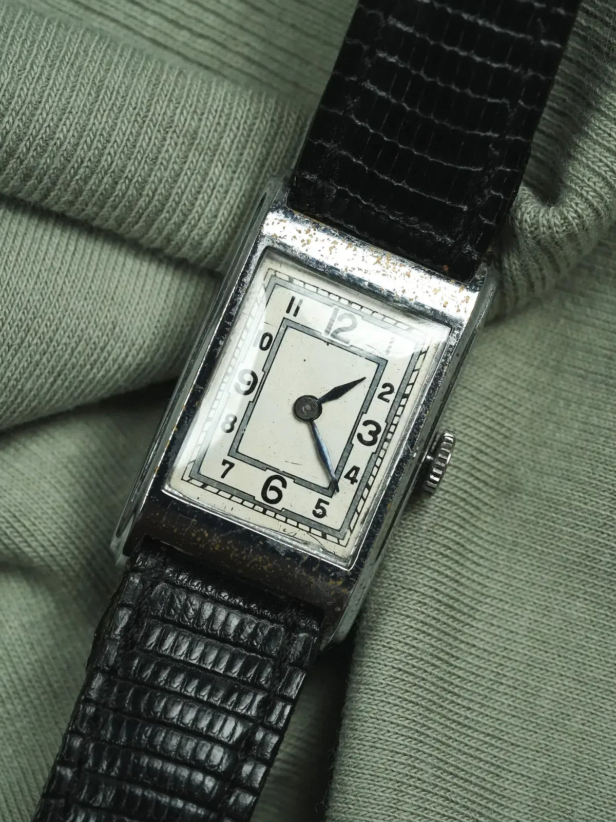 Vintage rectangular watch with white dial, black numerals, and textured black strap on green fabric