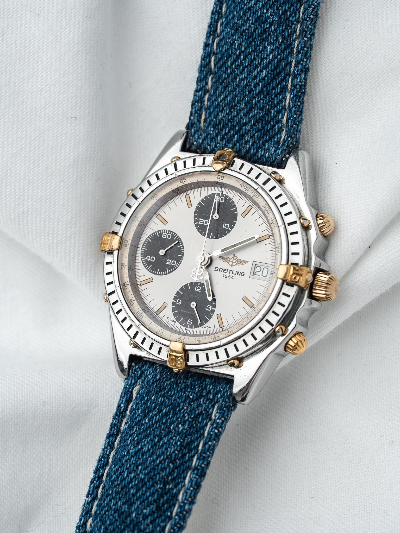 Breitling chronograph watch with silver dial, blue denim strap, gold accents on white background