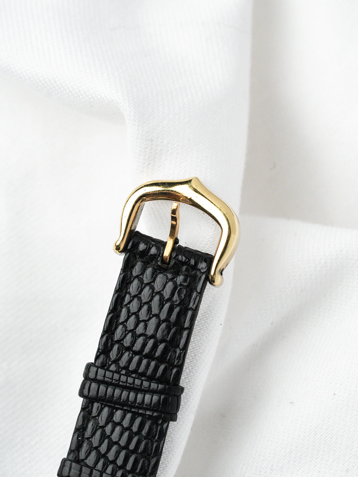 Black lizard skin watch strap with gold buckle on white fabric background