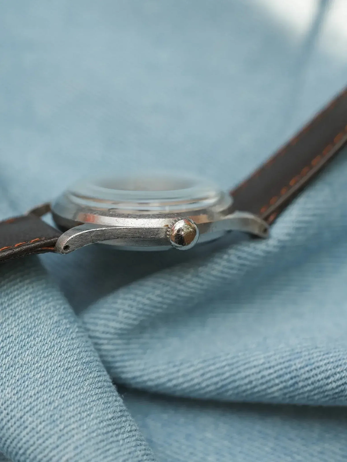 Vintage watch with steel case, brown leather strap, resting on denim fabric