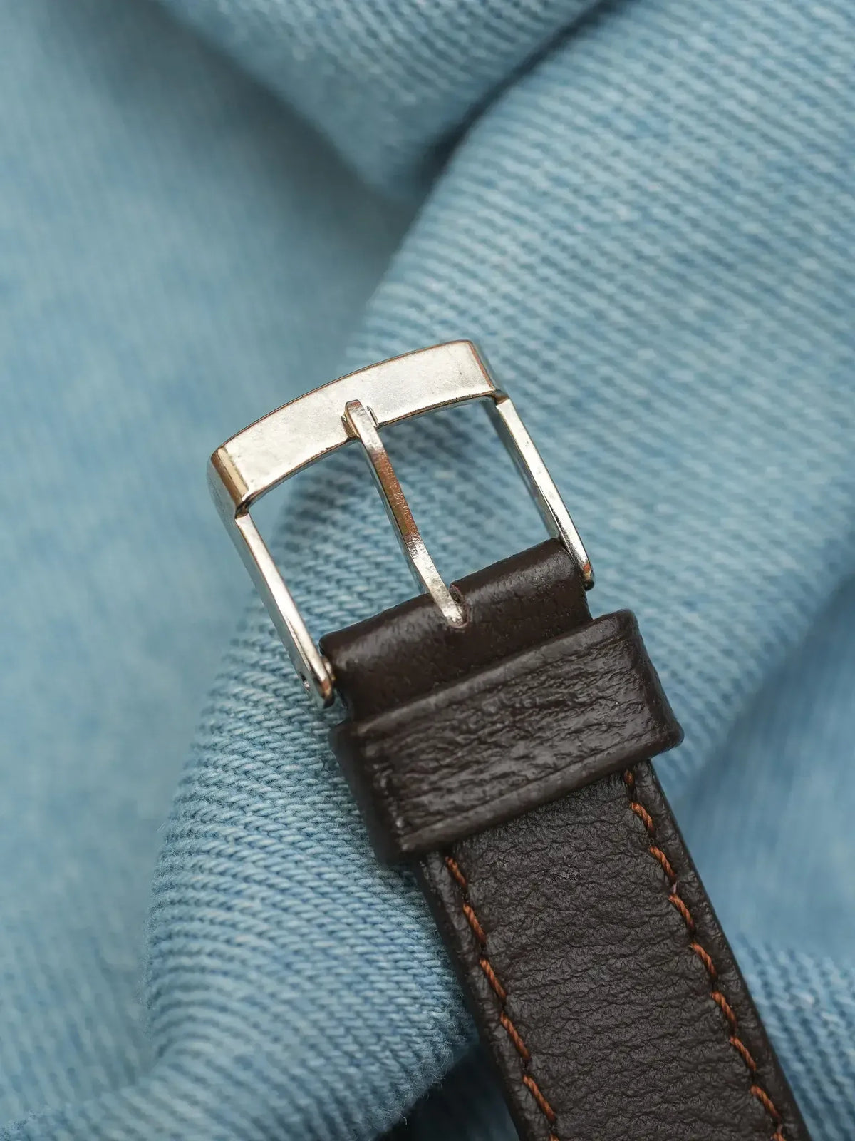 Brown leather watch strap with orange stitching and silver buckle on denim