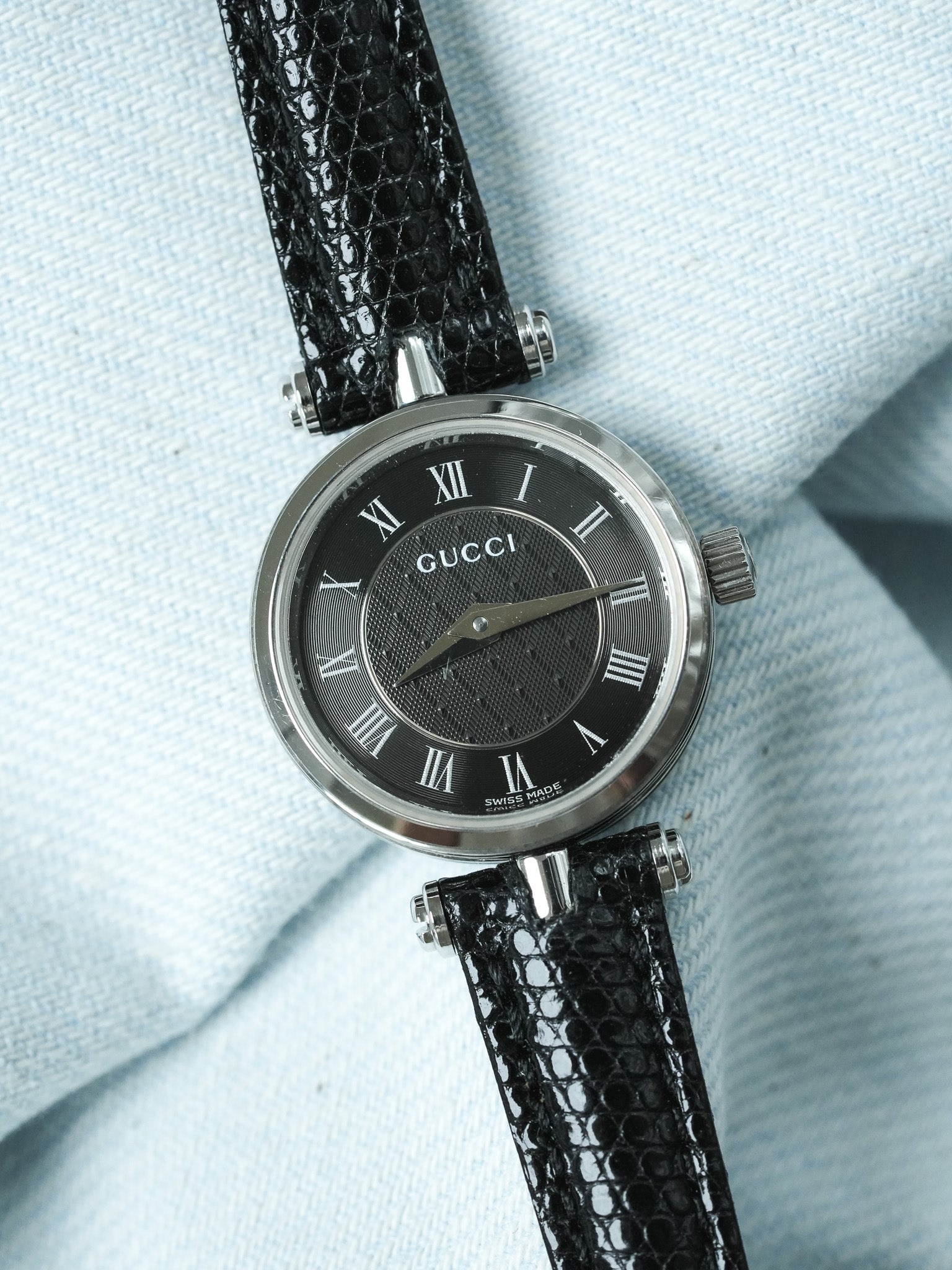 Gucci watch with black dial, Roman numerals, and black lizard strap on light fabric