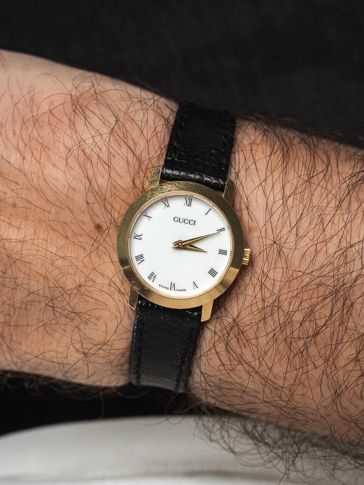 Gucci vintage watch with gold case, white dial, Roman numerals, black leather strap worn on wrist
