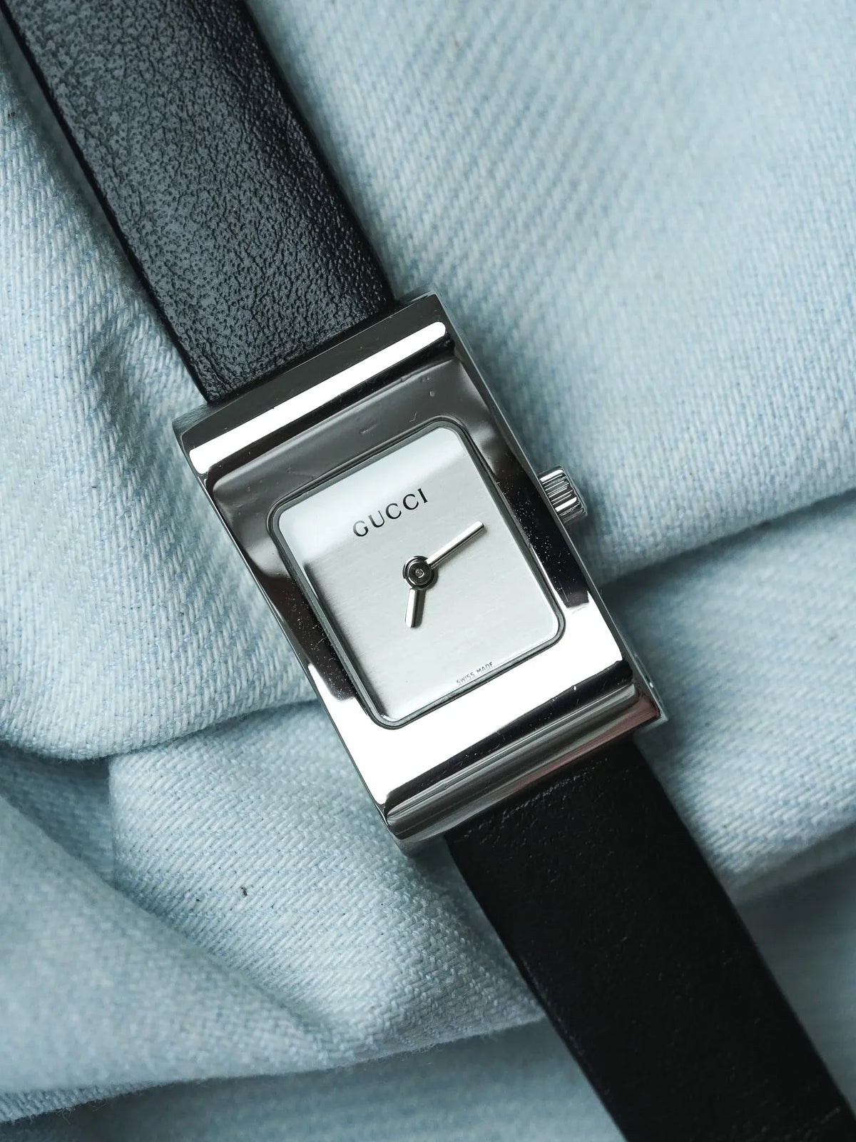Gucci vintage watch with rectangular silver case, white dial, black leather strap on light blue fabric