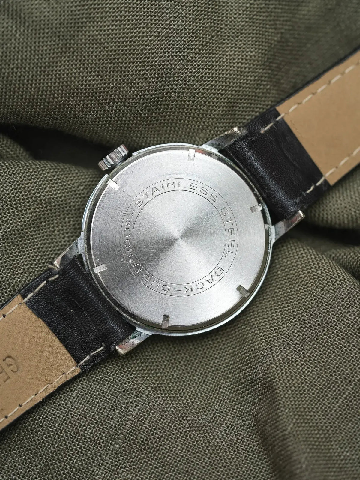 Vintage watch with stainless steel case back and black leather strap on olive fabric