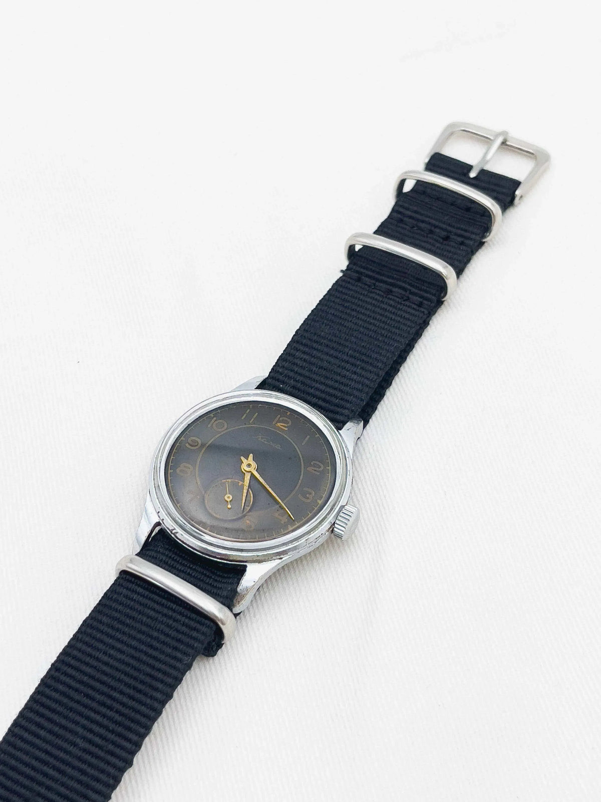 Vintage Tissot watch with dark dial, gold hands, steel case, black NATO strap on white background