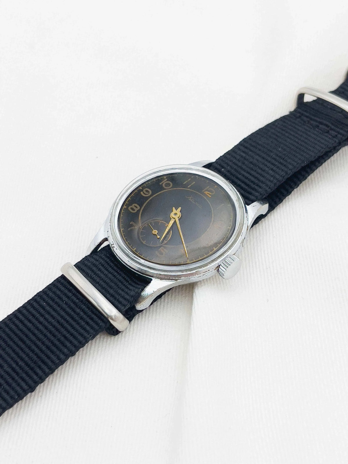 Vintage Universal Genève watch with black dial, gold hands, steel case, and black fabric strap