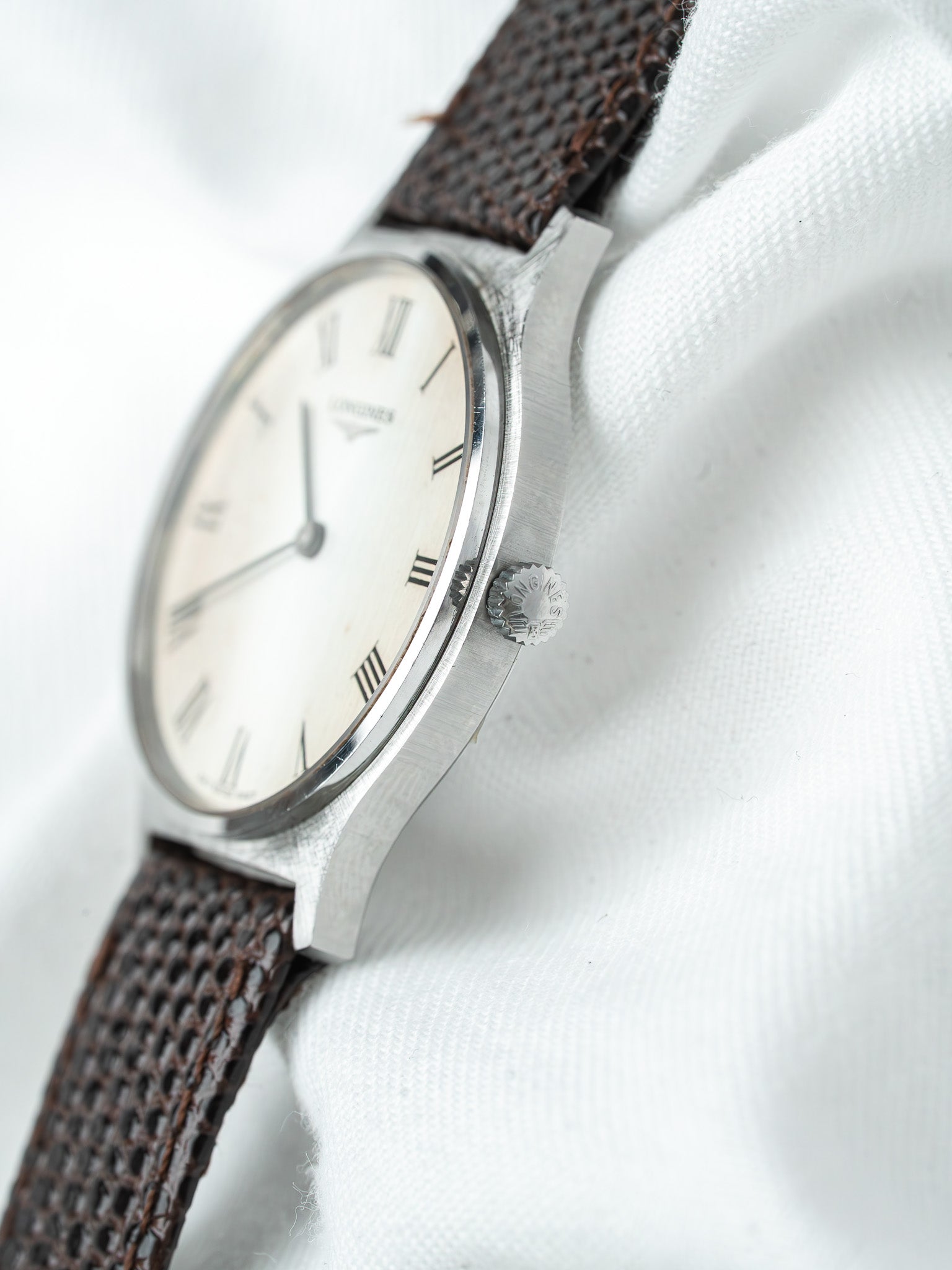 Longines vintage watch with white dial, Roman numerals, and brown textured strap on white fabric