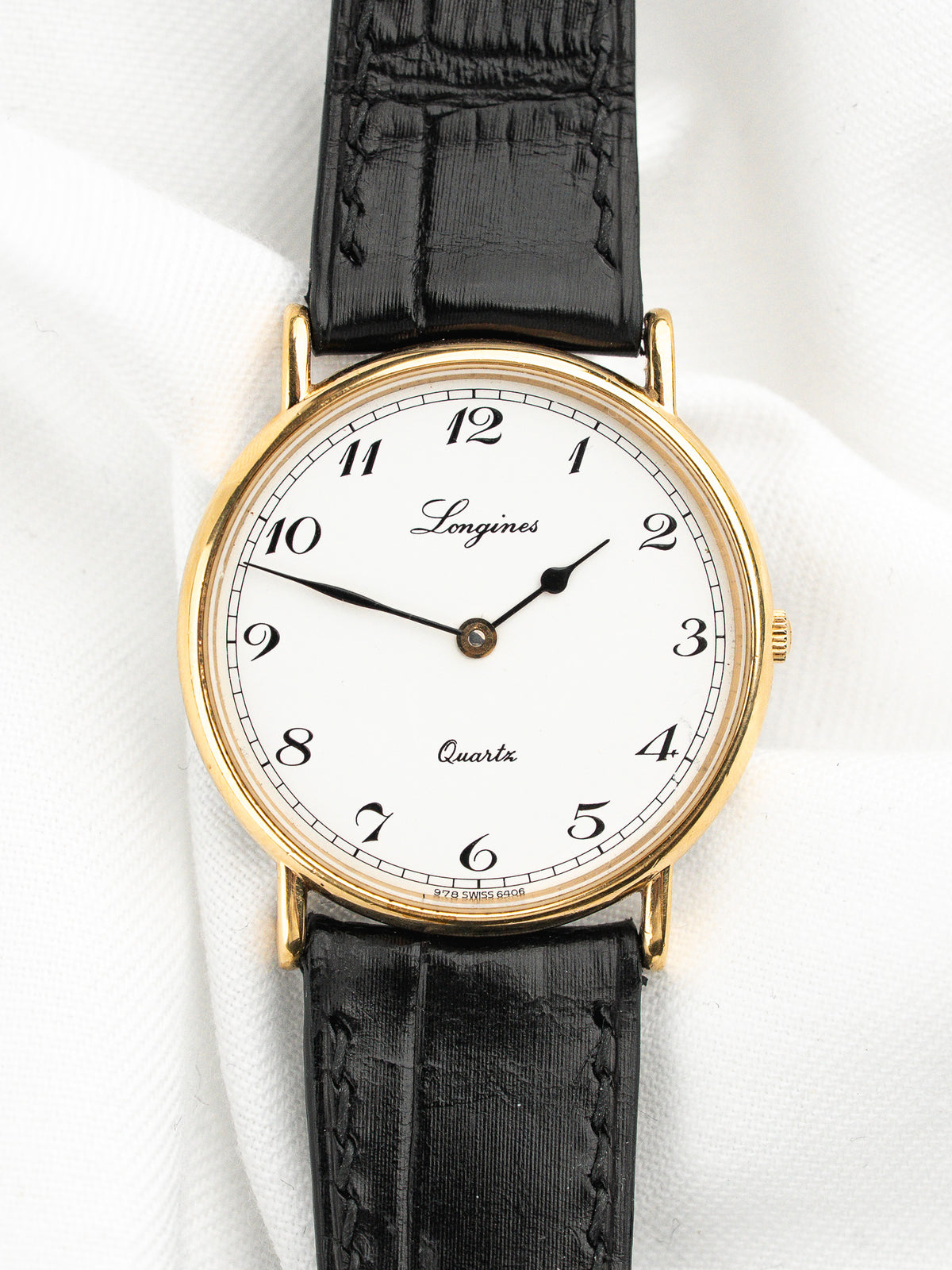 Montre Longines vintage au cadran blanc, boîtier doré et bracelet en cuir noir.