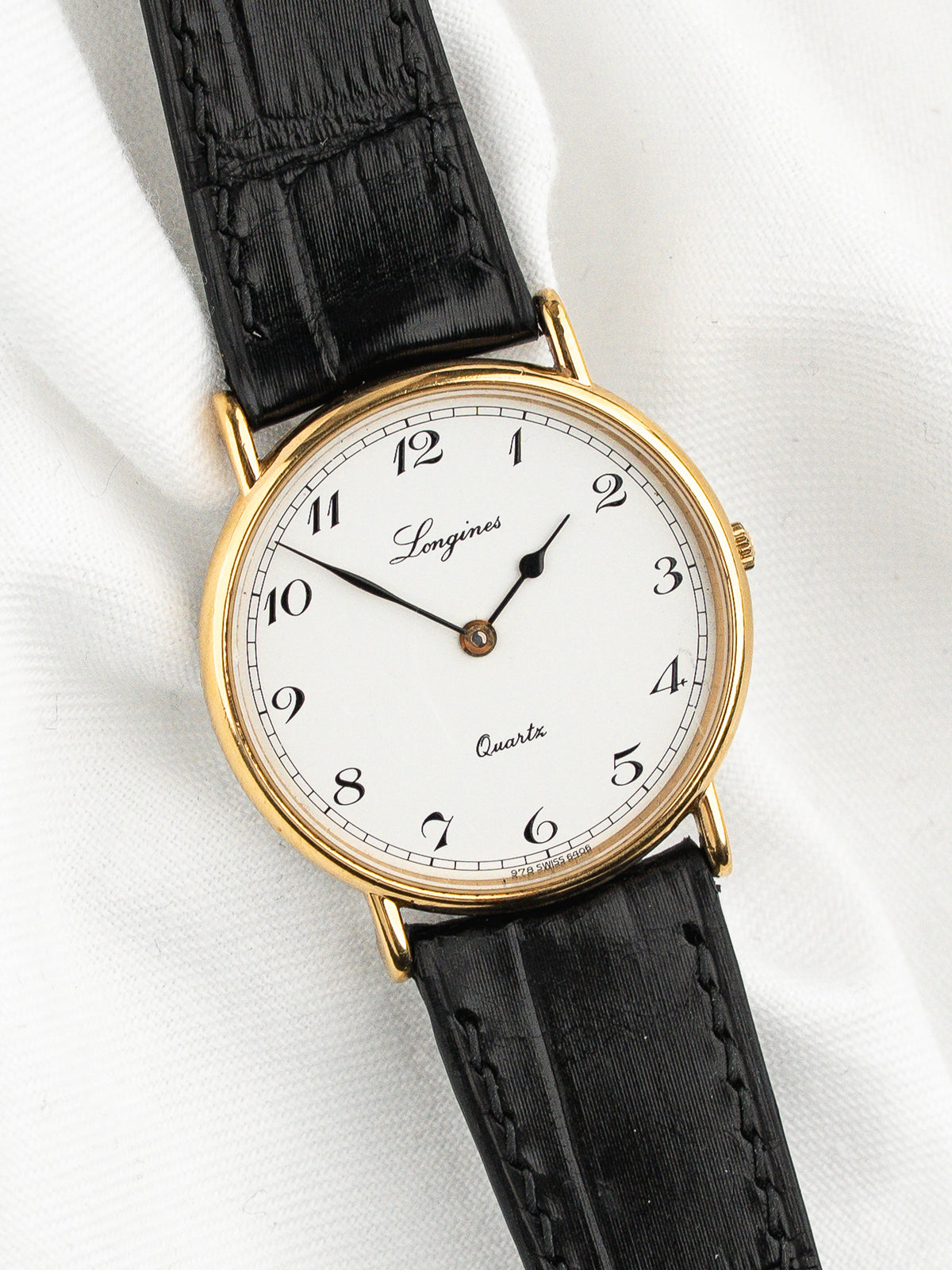 Montre Longines vintage au cadran blanc, boîtier doré et bracelet en cuir noir.