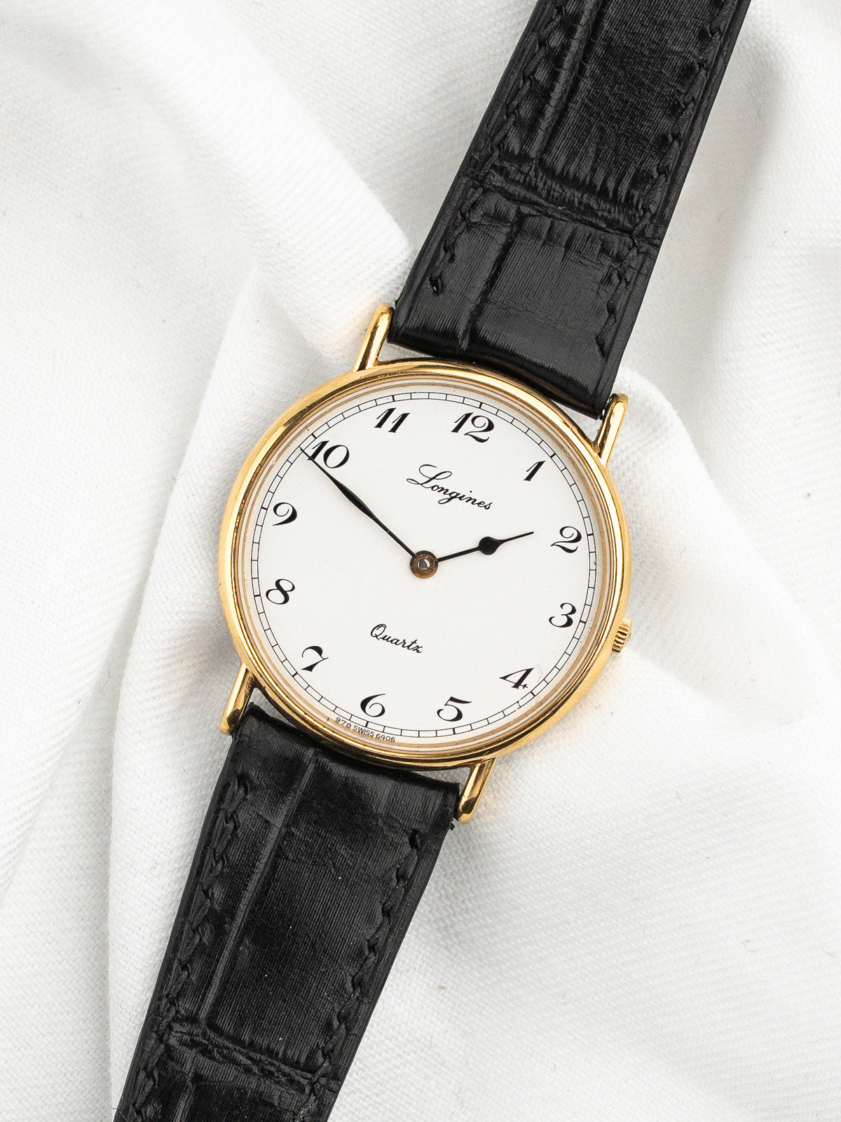Montre Longines vintage avec cadran blanc, boîtier doré et bracelet en cuir noir.