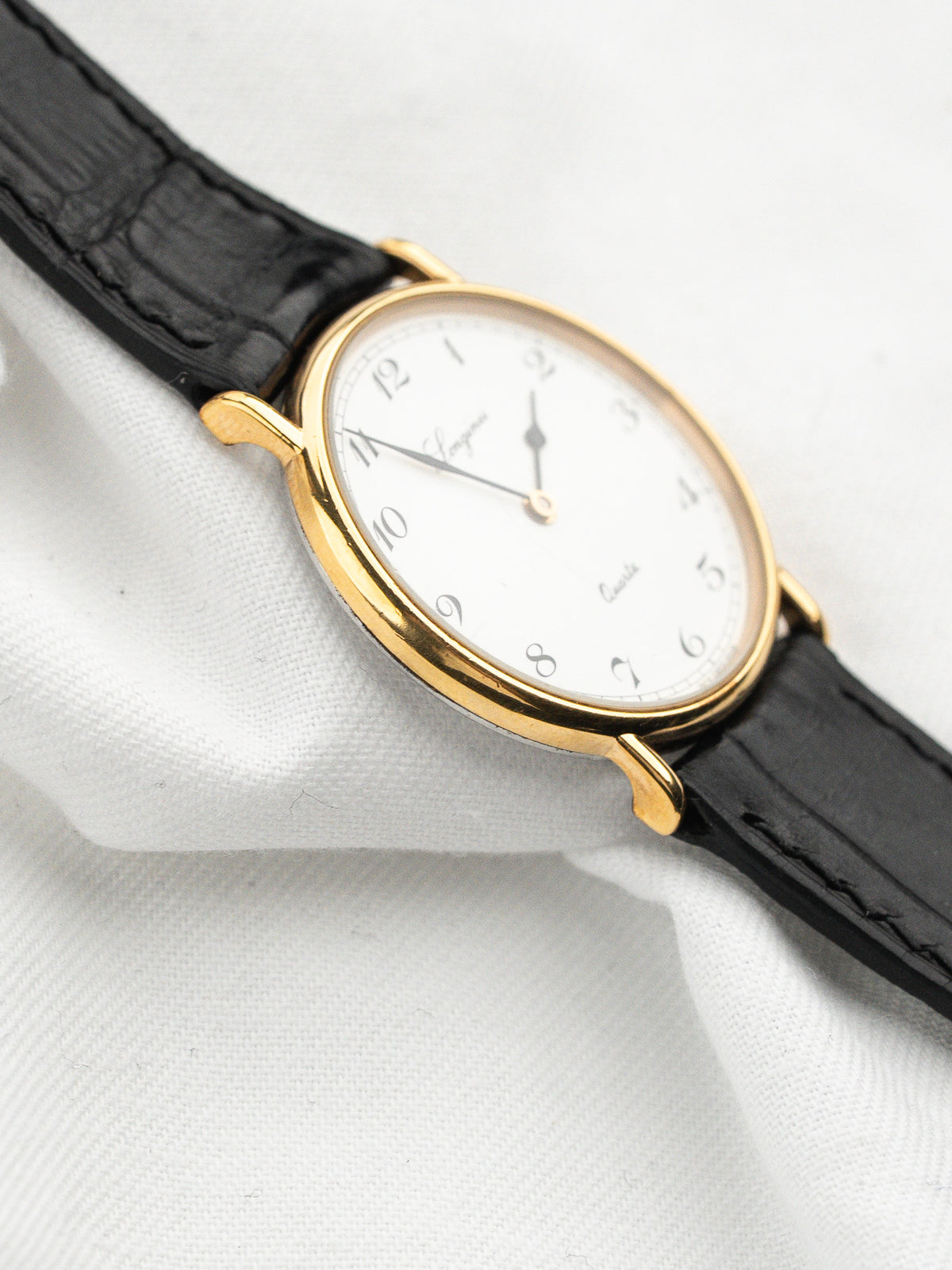 Montre vintage ovale avec cadran blanc, boîtier doré et bracelet en cuir noir.