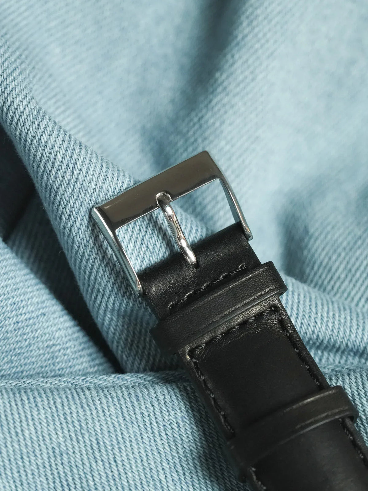 Black leather watch strap with polished steel buckle on denim background