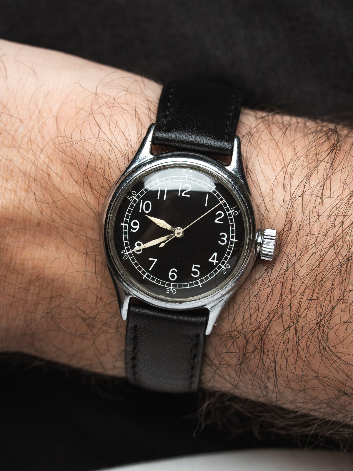 Vintage watch with black dial, white numerals, steel case, black leather strap on wrist