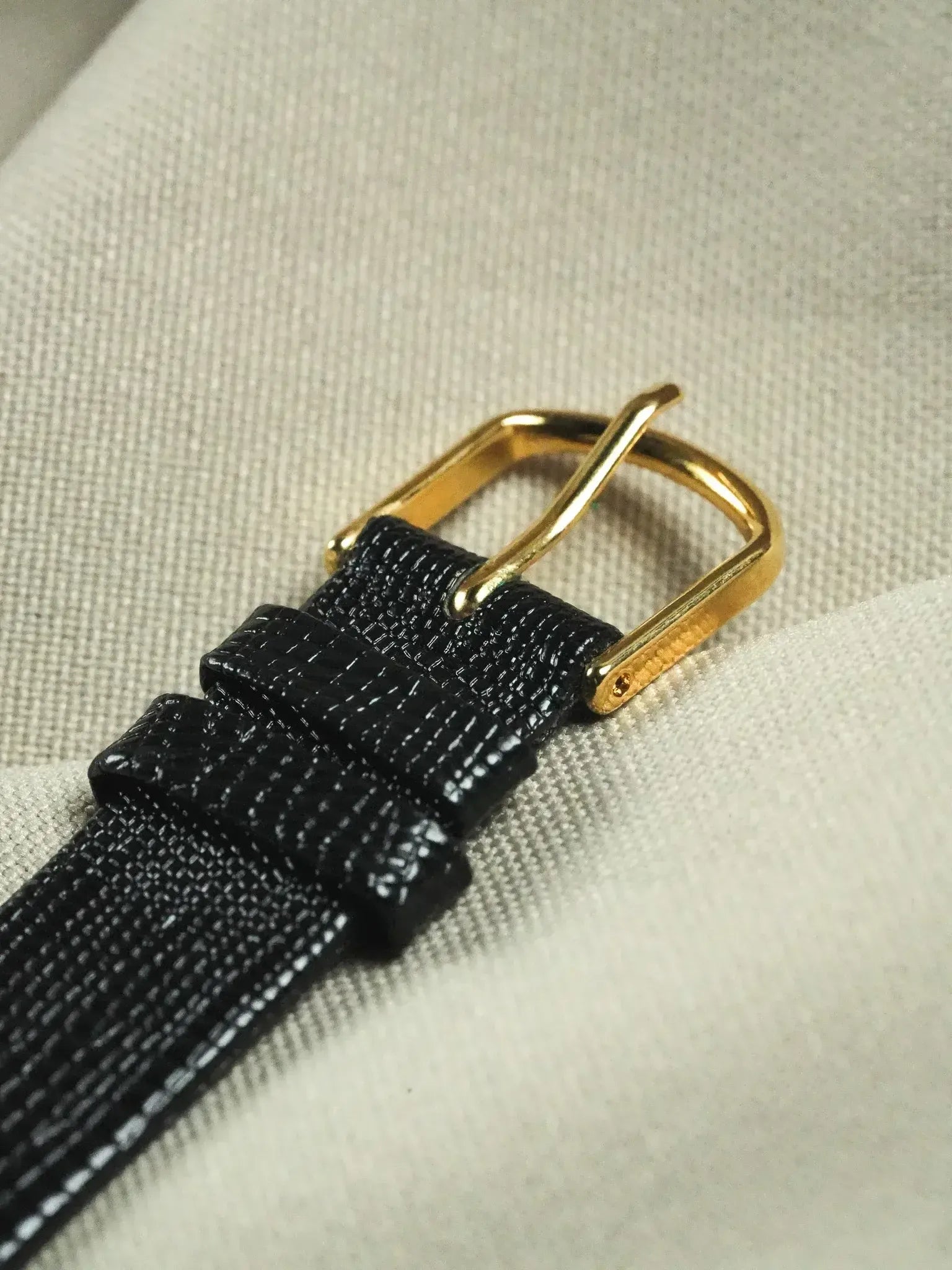 Black textured watch strap with gold buckle on neutral fabric background