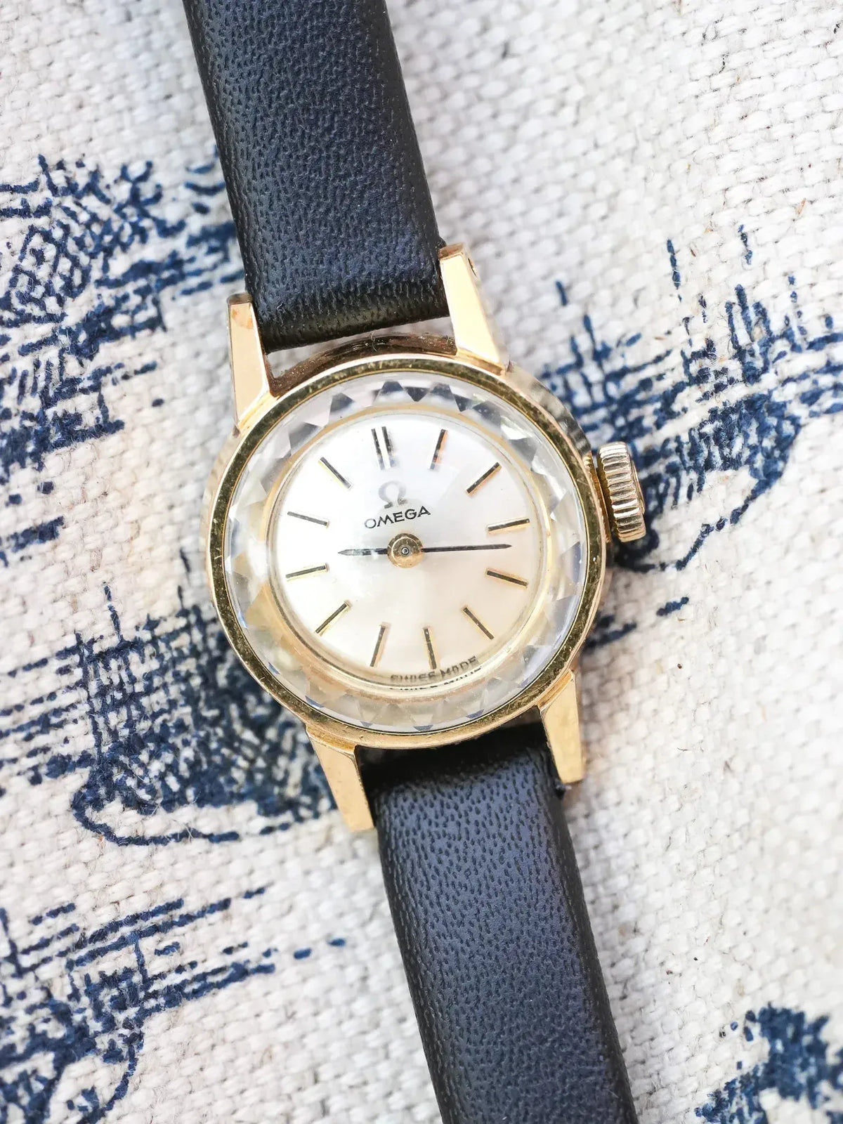 Vintage Omega watch with gold case, white dial, black leather strap on patterned fabric