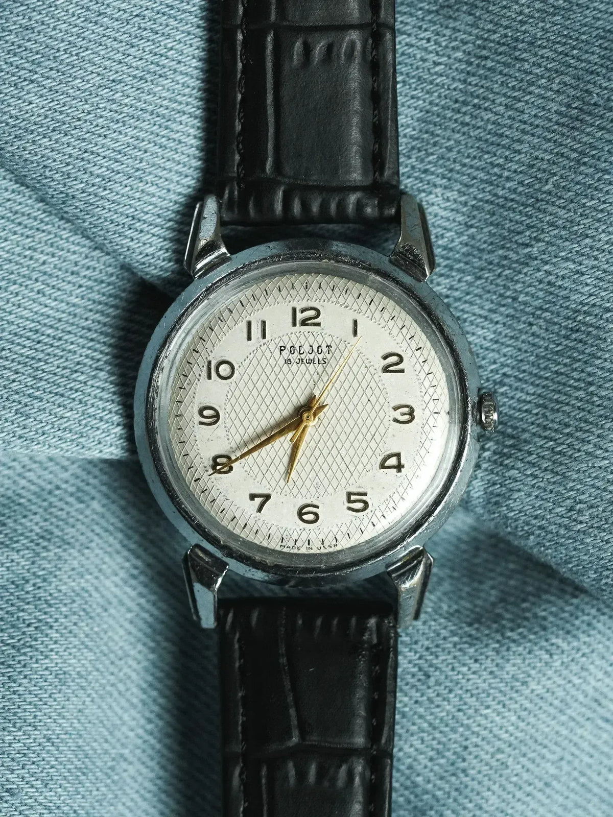 Vintage Poljot watch with silver dial, black leather strap, resting on denim